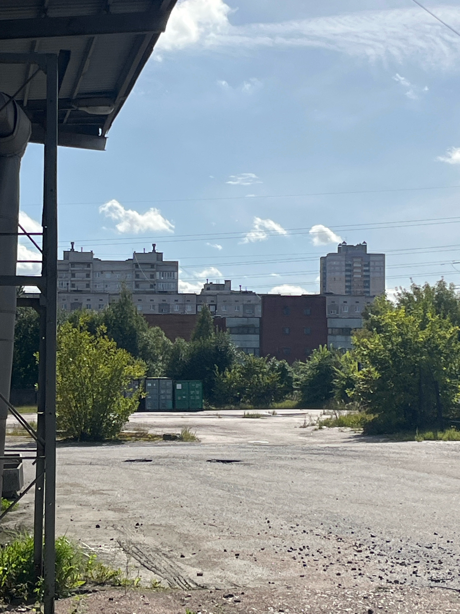 The image shows an outdoor scene in Saint Petersburg, Russia, on a sunny day with scattered clouds. In the foreground, a gray, gravelly asphalt surface is visible, with patches of grass and some debris. A dark gray metal structure, possibly part of a building or industrial equipment, is on the left side of the frame, obscuring some of the background.
In the middle ground, a line of lush green trees separates the foreground from a row of buildings. Behind the trees, several shipping containers, two green and one teal, are stacked.
The background features a series of multi-story apartment buildings, characteristic of Soviet-era architecture, with light gray facades and numerous windows. The tallest building is a light-colored, rectangular structure with visible window details. Overhead, power lines crisscross the sky. No people are visible in the image. The overall impression is one of an urban industrial or residential area.