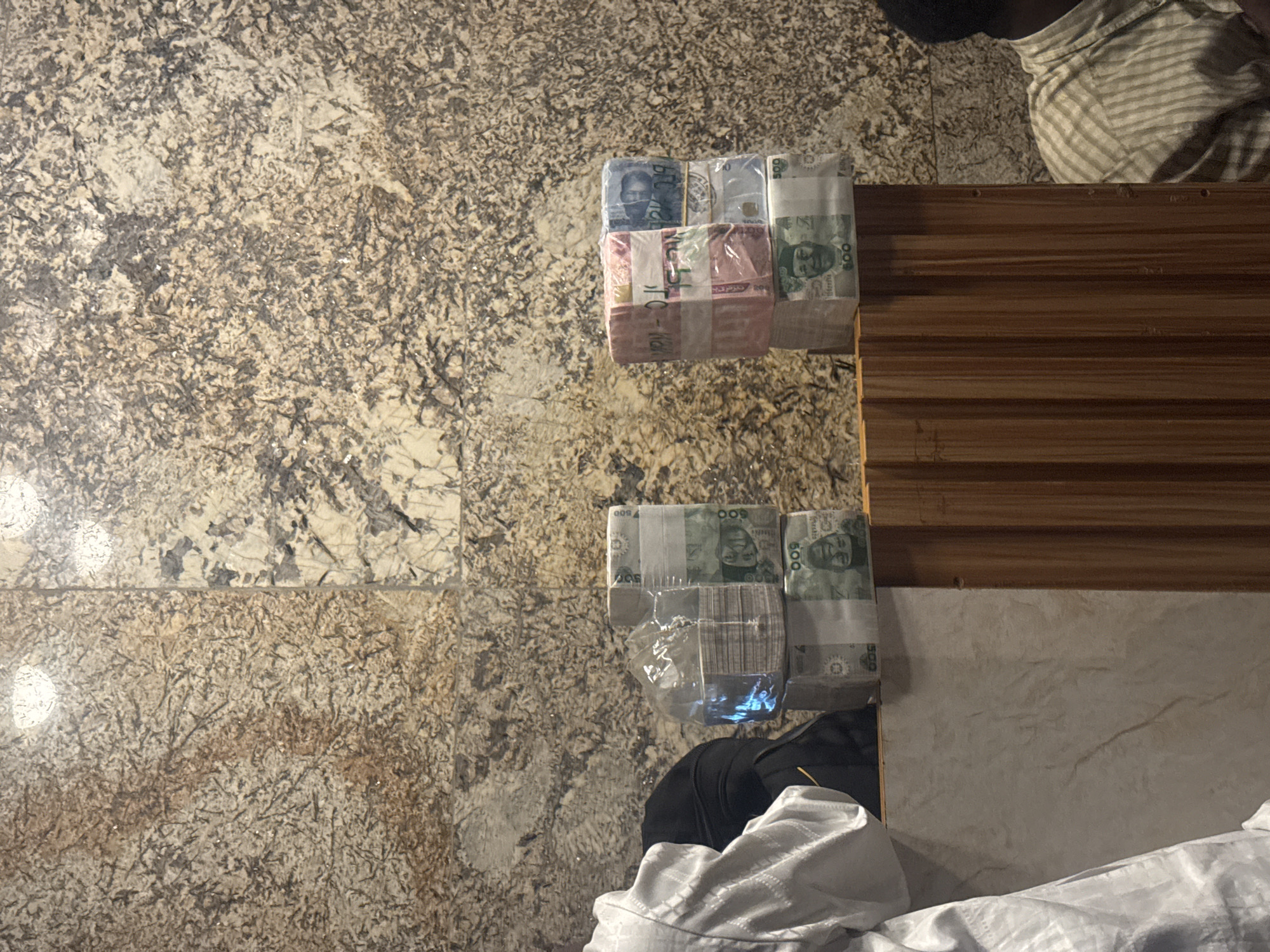 In Abuja, Nigeria, a person wearing a white garment stands to the left of the frame, partially obscuring stacks of Nigerian Naira currency. Two distinct piles of banknotes are visible, each wrapped in plastic. The stack on the left is a tall, thin bundle of what appears to be 500 Naira notes, each bound with a white band and plastic wrap. To the right, there are two stacks of banknotes. The lower stack also consists of 500 Naira notes, similar to the left stack. The upper stack is composed of colorful banknotes, with some appearing pink and others blue, also bound with white bands and plastic wrap. The currency is placed on a marbled surface, which transitions to a wooden surface in the foreground. The background features a textured, mottled wall. Another individual, wearing a striped shirt, is visible on the right side of the frame, partially obscured by the wooden surface. The lighting suggests an indoor setting.