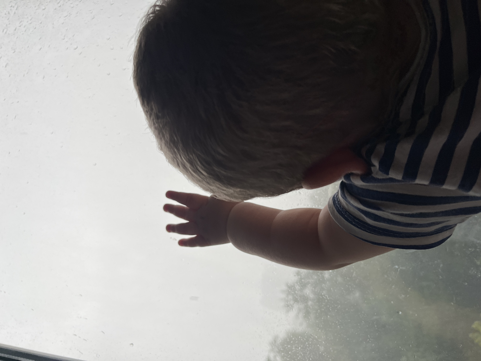 A child's hand, with fingers splayed, is pressed against a rain-streaked window. The child's head and shoulder are visible in the foreground, with light-colored hair and a blue and white striped shirt. The background is obscured by the rain and mist on the window, but hints of green foliage can be seen through the glass. The scene is overcast and appears to be indoors looking out. The image does not provide any information to determine its location in Chelyabinsk, Russia.