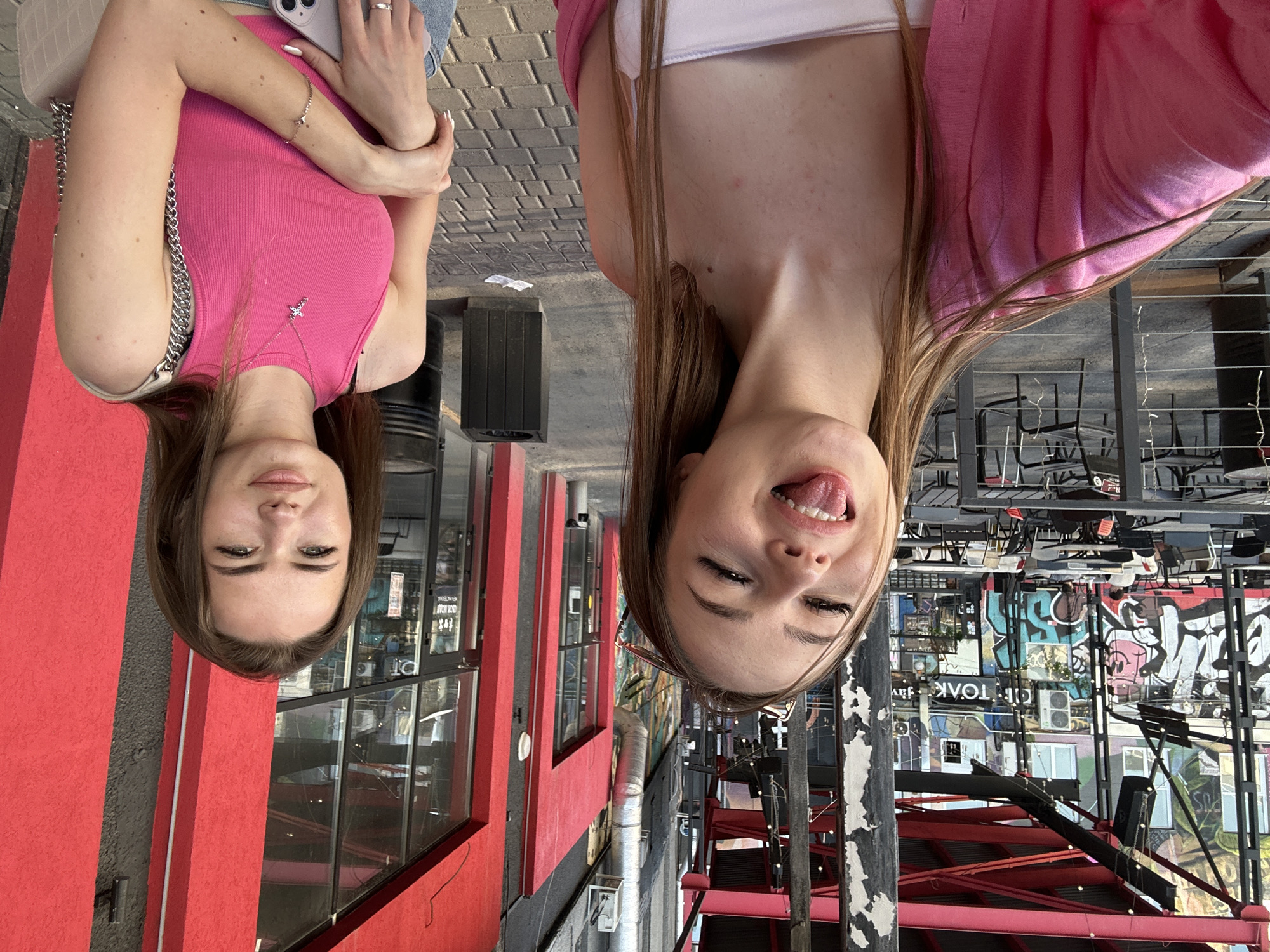 Two young women are depicted in an outdoor urban setting, likely posing for a casual photograph or selfie. The image is oriented vertically with the subjects' heads pointing towards the bottom.
The woman on the left wears a pink ribbed tank top, with her arms crossed and a light-colored mobile phone clutched in her right hand. She accessorizes with a silver necklace featuring a cross pendant, a silver bracelet, and a silver chain crossbody bag. Her long brown hair frames a face with a neutral, calm expression, and her eyes are open.
The woman on the right, positioned slightly behind the first, is more playful. She wears a pink top over a white garment and has long brown hair. Her eyes are partially closed, and she is sticking out her tongue, conveying a lighthearted mood.
The background is an urban landscape, suggesting a city street or a commercial district. To the left, a vibrant red building facade with windows is visible, reflecting some indoor elements. The right side of the background features a more industrial or eclectic aesthetic with exposed structures, possibly part of an outdoor dining area, indicated by numerous dark chairs and tables. Walls covered in colorful graffiti are prominent, with the word "TOAK" clearly visible on a white rectangular sign amidst the artwork. The overall lighting suggests it is daytime, with bright conditions.