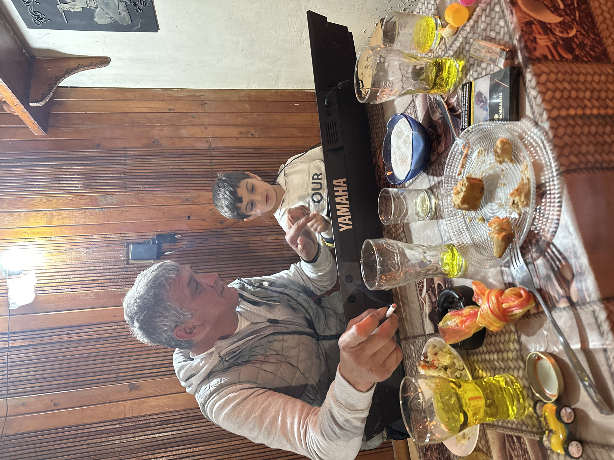 This indoor scene, captured in Mlekarevo, Bulgaria, depicts an older man and a young boy seated at a table laden with food and drinks, alongside an electronic keyboard. The image is oriented sideways, featuring a warm, wood-paneled wall on the left and a lighter-toned ceiling above.
On the left, an older man with short grey hair and a light-colored hooded jacket holds a lit cigarette in his right hand. He is looking towards the young boy. The boy, with dark hair, is to the man's right, wearing a white hoodie with the word "OUR" printed on it. He appears engaged with a black Yamaha electronic keyboard positioned on the table between them, with his hands near the keys. The brand "YAMAHA" is prominently displayed on the instrument.
The table is covered with a patterned brown and white tablecloth. Scattered across it are multiple clear glasses, some containing a yellow liquid. A clear glass plate holds several pieces of partially eaten pastries or sweets. Another plate contains a colorful dish, possibly a salad, alongside black olives. A blue bowl filled with a white granular substance (likely sugar), a pack of cigarettes (faintly showing "MARLBORO" branding), a lighter, a fork, a knife, and a small yellow toy car are also visible on the table. A decorative wooden shelf and a dark rectangular wall hanging with a light-colored relief sculpture are mounted on the wall above the wooden paneling.
The scene suggests a casual and relaxed family gathering, possibly during an evening or late afternoon, given the indoor lighting. The boy seems curious and engaged with the keyboard.