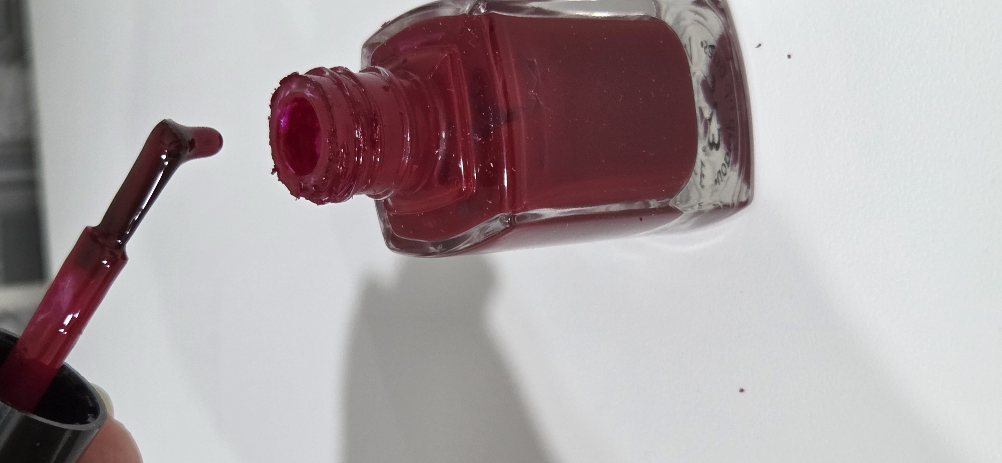The image presents a close-up, slightly elevated perspective of an open, clear glass nail polish bottle filled with a dark red, viscous liquid. Above the bottle, a nail polish brush, attached to a black handle, is positioned, actively dripping a single drop of the red polish downward towards the bottle's opening. The brush head is fully saturated with the deep red substance. The bottle's neck is visibly caked with dried or smeared red polish residue, and faint, inverted text or numbering, such as "3" and "MOD", is visible on the bottom of the glass bottle. The scene is set on a uniform white surface, likely a tabletop, exhibiting soft, indistinct shadows on the left side. Small, dried specks of red polish are scattered on the white surface near the base of the bottle. No individuals are directly visible in the frame, but the action implies a person holding the brush. The image contains no visual elements or contextual information that would identify its location as the city of Lipetsk, Russia, or any other specific geographical region.