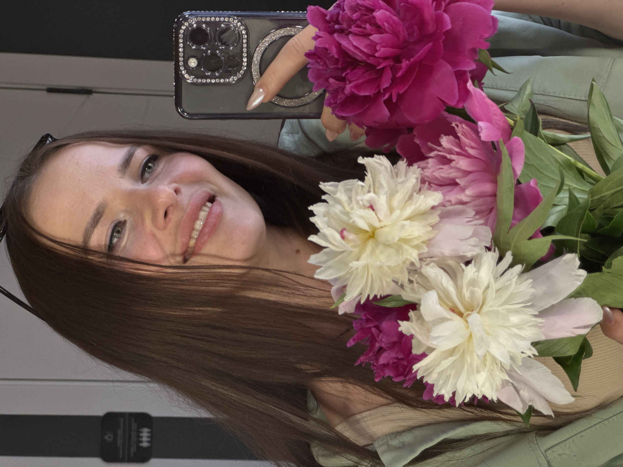 A young woman is taking a selfie in a mirror. She is holding a bouquet of pink and white peonies in front of her. She is wearing a light green jacket over a beige top. Her hair is long and brown, and she has braces on her teeth. The background appears to be an indoor setting, possibly a room, with a grey and black wall on the left and a lighter wall on the right. The woman's phone is visible in her hand, and the phone's case is decorated with rhinestones. The image does not contain any information that would allow for its location to be identified as Chaikovsky, Russia.