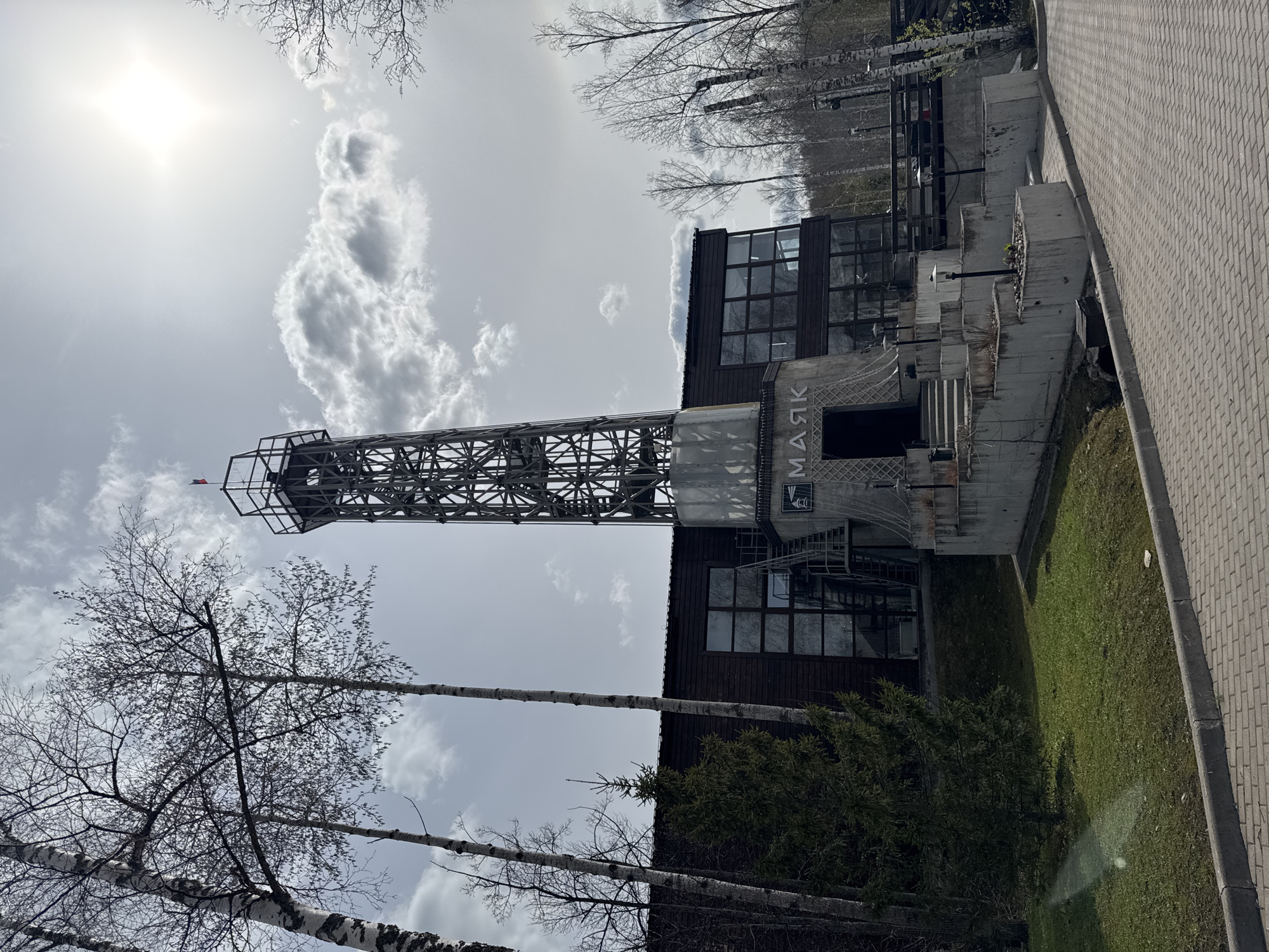 The image depicts a view of the "Mayak" art park in the city of Pavlovka, Russia. A lattice tower stands adjacent to a modern building featuring large windows and concrete elements. Bare trees are visible in the foreground and against a cloudy sky. The surrounding landscape includes a grassy area and a paved walkway.