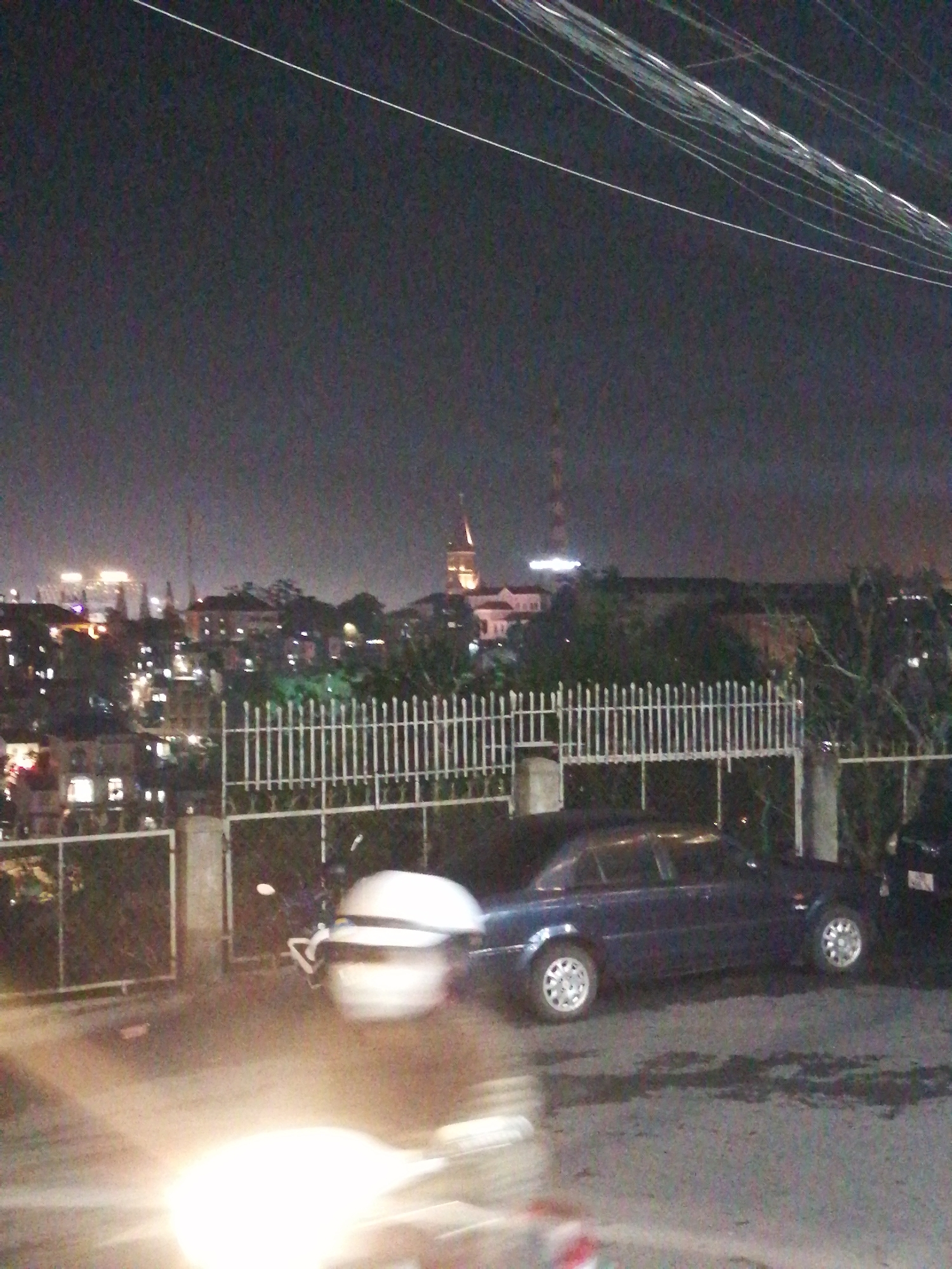 The image depicts a nighttime street scene in Da Lat, Vietnam. A dark blue sedan is parked along a road behind a white metal fence. A white scooter is parked near the sedan, with only part of it being visible. In the background, the cityscape of Da Lat is visible, with several buildings, including a structure with a spire and a radio tower. Streetlights and building lights illuminate the city. Overhead, a cluster of electrical wires stretch across the sky. The image appears to be taken from a low angle, capturing the tail lights of a motorcycle or scooter in the foreground.
