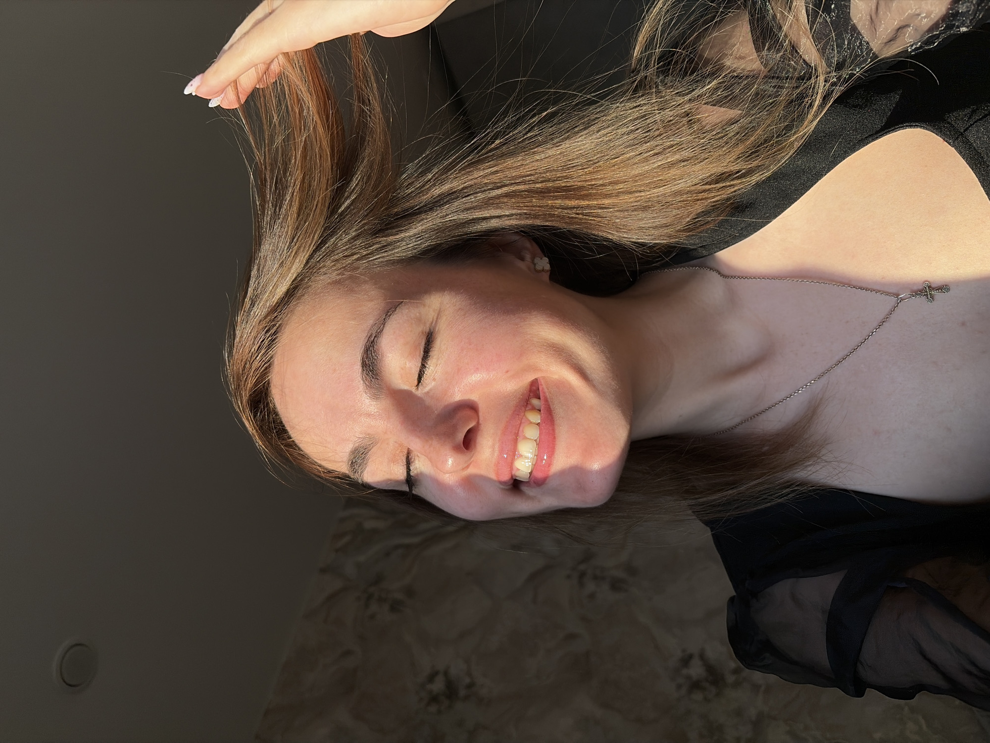 A young woman with long, light brown hair is smiling with her eyes closed. She is wearing a black top and a silver necklace with a cross pendant. Her hand is raised, touching her hair. The background appears to be a dark room or area with varying levels of light. The image does not provide specific information to place the woman in Izhevsk, Russia.