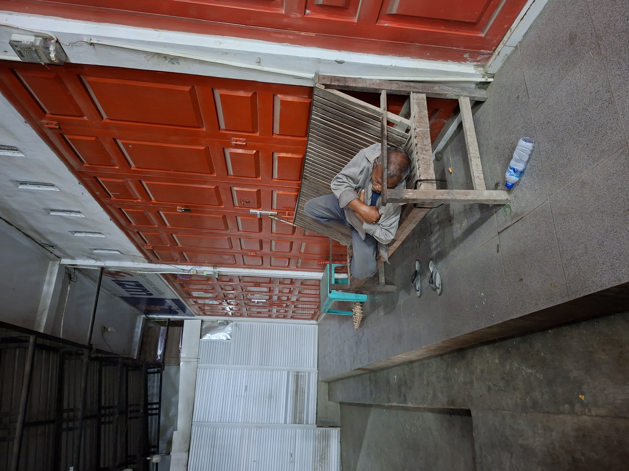 A man sits on a wooden structure next to a red garage door in what appears to be Keudegeudong, Indonesia. He is wearing a grey shirt and pants. A clear plastic water bottle lies on the grey tiled floor next to him. The background includes a white metal fence and black metal racking. A turquoise plastic chair is next to the wooden structure. The scene is rotated for viewing perspective.