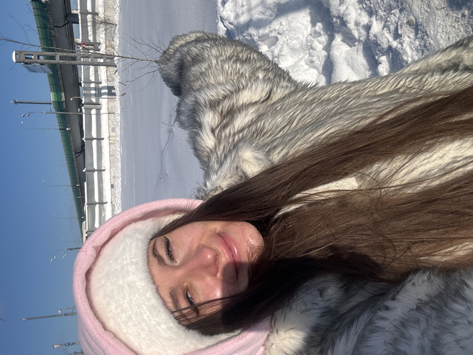 The image displays a young adult female in a close-up, selfie-style photograph taken outdoors in Shul'gino, Russia, on a clear, sunny winter day. The individual is positioned slightly to the left of the frame, smiling faintly with open eyes and visible eyelashes. She wears a pink hood over a white, fluffy hat and a thick, light grey and white faux fur coat. Her long, dark brown hair cascades over her right shoulder and chest, casting a shadow on her left cheek. Her right arm, covered by the fur coat, is extended towards the right side of the frame, suggesting it holds the camera. The background consists of a broad, snow-covered landscape. A small, bare sapling protrudes from the snow in the mid-ground to the right. Further back, an elevated multi-lane highway or bridge, featuring a green barrier along its edge, is supported by concrete pillars. Concrete retaining walls, partially covered in snow, are visible beneath the elevated structure, and streetlights line the highway. The sky is clear blue.