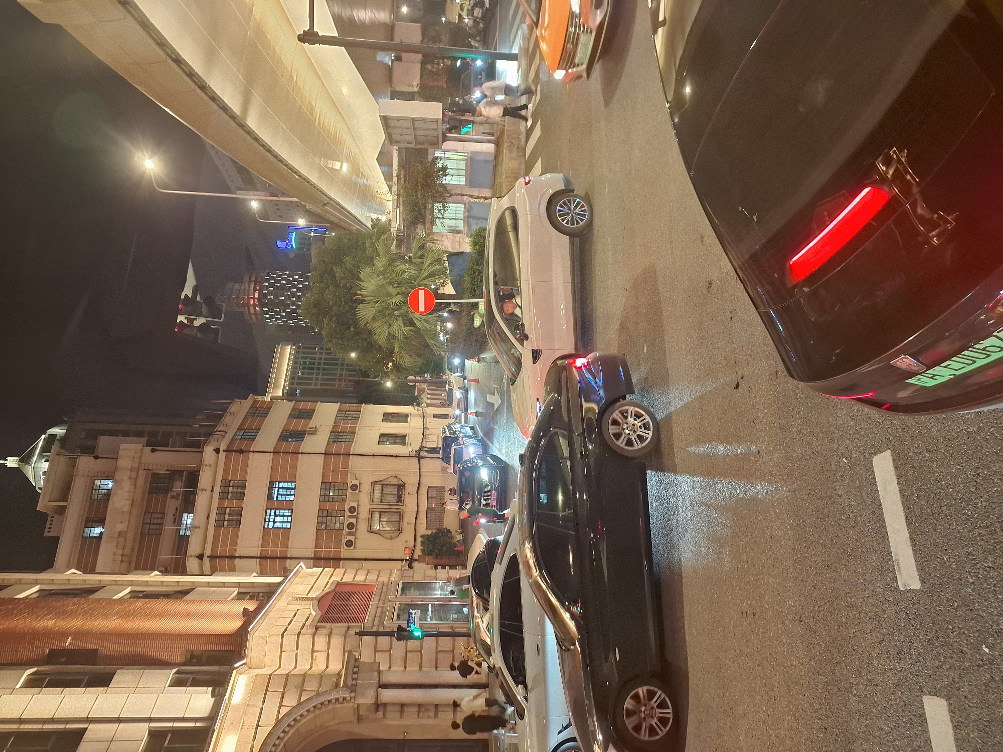 The image depicts a nighttime street scene in Shanghai, China, characterized by a mix of older and modern architecture, traffic, and illuminated signs. A multi-lane road is occupied by several vehicles, including a black BMW sedan in the foreground, a white sedan, a light-colored SUV, and an orange car. Pedestrians are visible on the sidewalk and crossing the street. The environment includes traffic lights, street lamps, and elevated roadways, suggesting an urban setting. Buildings on either side of the street exhibit architectural styles ranging from classical to contemporary, with lights illuminating various windows. The overall impression is one of a bustling city at night, with the flow of traffic and the presence of diverse structures.