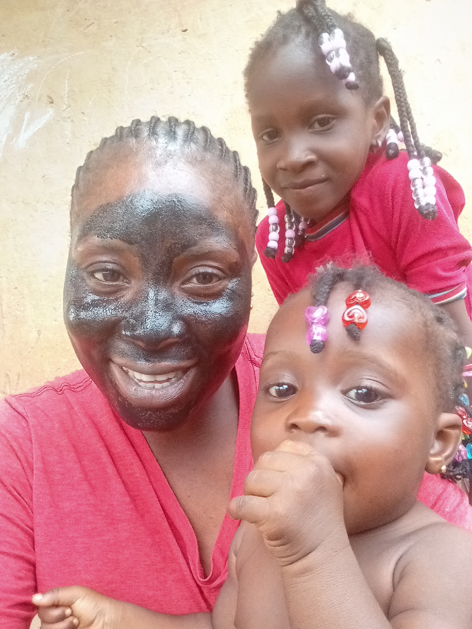 The image shows a woman and two children. The woman is in the foreground and has a dark substance applied to her face. She is smiling and wearing a red t-shirt. One child is behind her, wearing a red top and with braids adorned with beads. The second child is closer to the camera, with their hand near their mouth, and beads in their hair. The background is a plain, light-colored wall. The location is Akwanga, Nigeria.