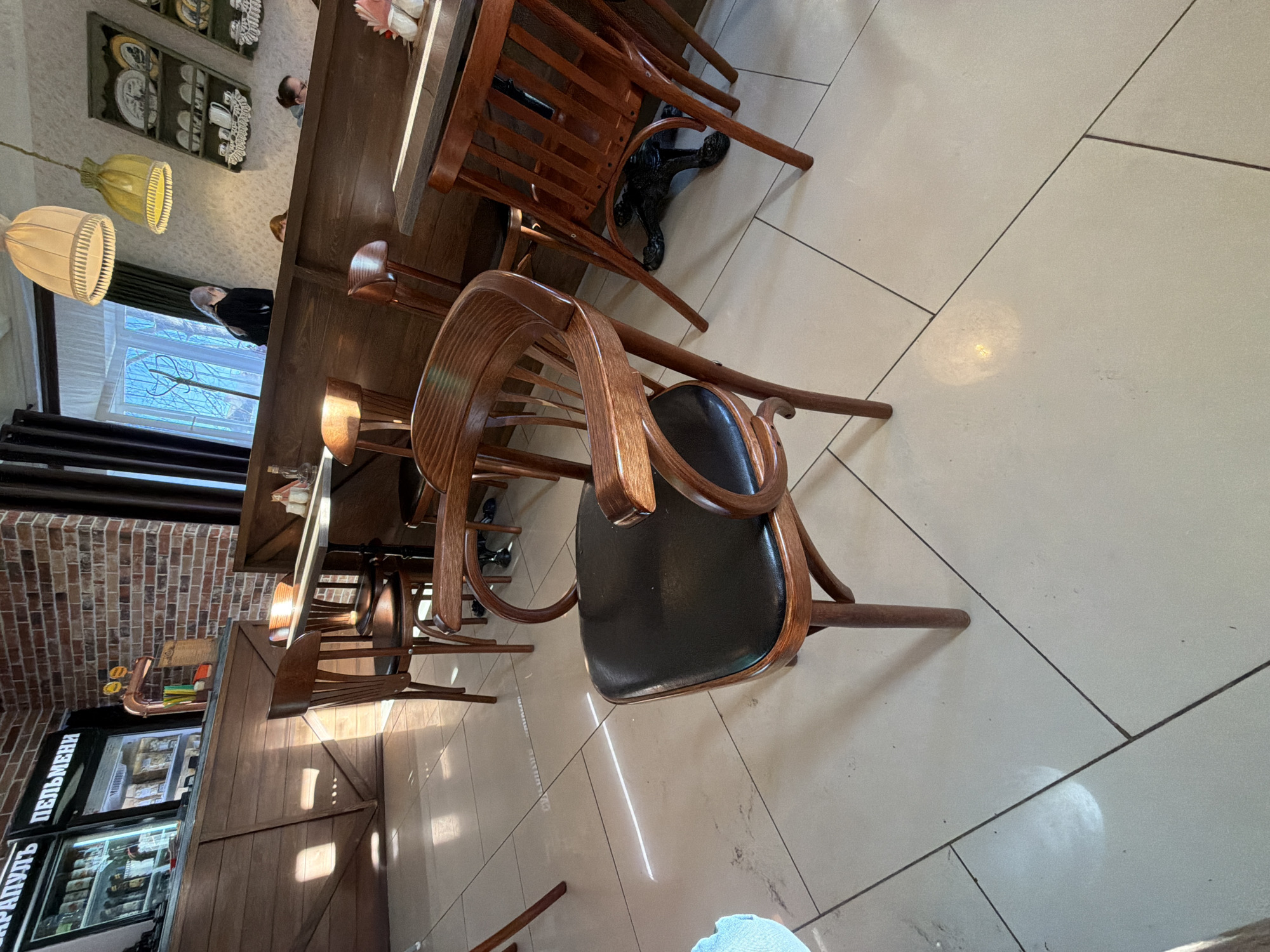 The image captures a restaurant interior. The focus is on a dark wooden chair with a black leather seat, positioned on a tiled floor with light-colored tiles. In the background, a wooden table is visible, surrounded by more chairs. A partially visible bar area includes a refrigerated display case with Cyrillic text ("пельмени" and "сарапуль"). The restaurant is illuminated by hanging lamps. The brick wall on the left side and the window area with a view to the outside are also visible. The perspective is angled, looking slightly downward towards the chair. The location is identified as Sarapul, Russia.