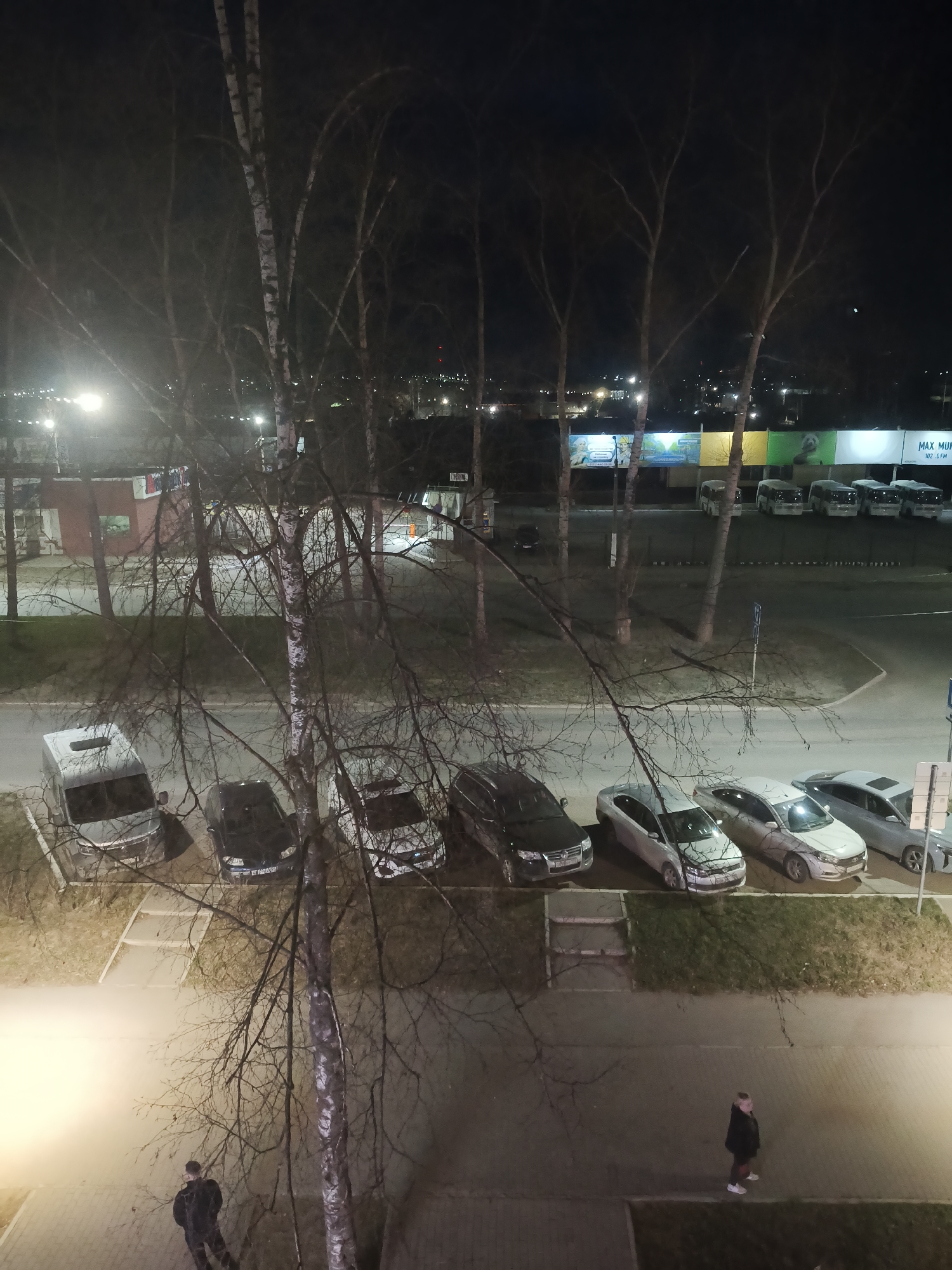 The image shows a nighttime view from an elevated position, likely a window. The scene overlooks a street in Chaikovsky, Russia with parked cars and trees. Six parked cars are visible, predominantly silver and white, along with a dark SUV. In the background, there are vans parked in front of a building with large advertisements. The presence of birch trees suggests the local flora. The street is illuminated by streetlights. Two individuals are walking on the sidewalk below. A man is walking towards the right of the image, another man walking toward the left.