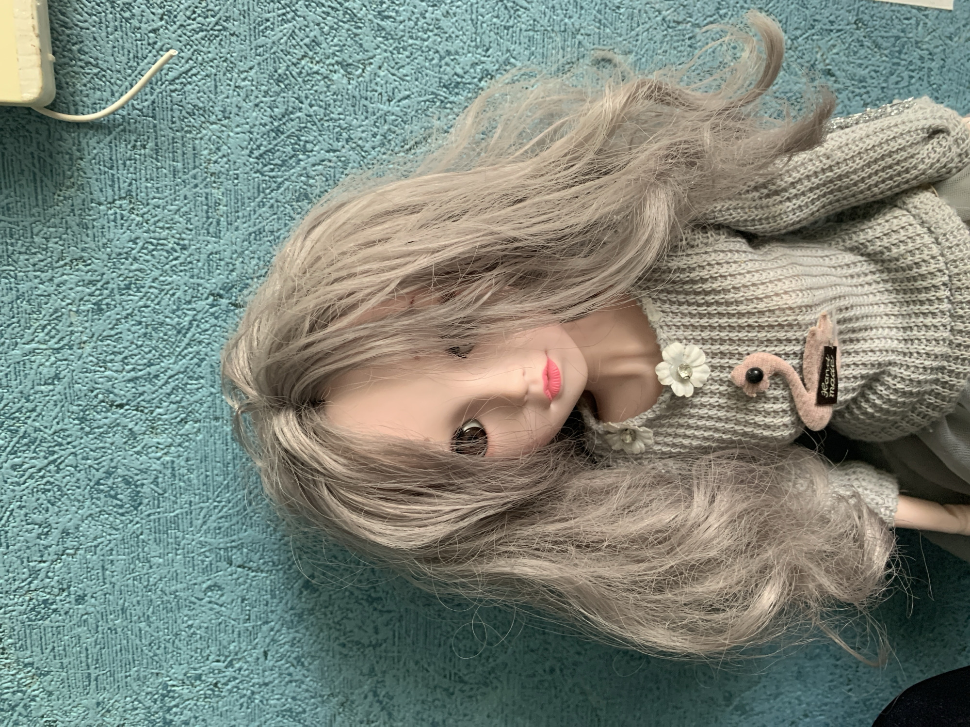 This media file features a doll with long, wavy, light gray hair and brown eyes, lying on a textured blue surface that resembles a carpet or mat. The doll is wearing a gray knitted sweater adorned with a white flower and a pink flamingo-shaped appliqué. A small tag with black text is attached to the sweater. The doll's head is tilted to the side, and its pink lips are slightly parted. To the upper left of the image, a white wire emerges from a white rectangular object. The overall scene is a close-up shot of the doll, with the blue textured surface filling the background. There are no indications of time of day, weather, or a specific event taking place. The location context is Volzhsky, Russia.