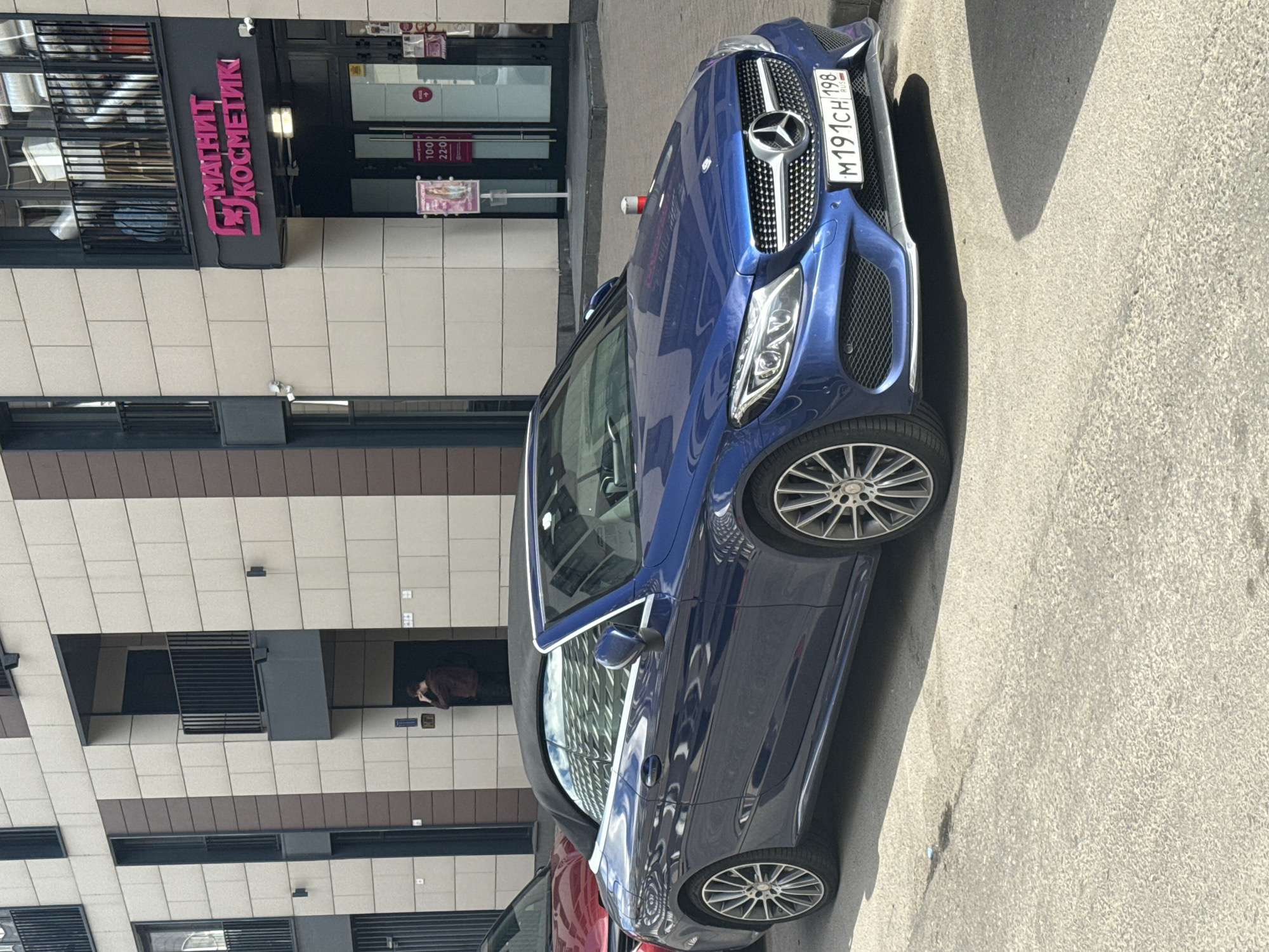 This image captures a sunlit street scene in Saint Petersburg, Russia, featuring a blue Mercedes-Benz convertible parked in front of a building with a "Magnit Kosmetik" store. The store's facade is marked with pink Cyrillic lettering, and a sign inside indicates operating hours of "10:00 22:00". The car's license plate reads "M 191 CH 198 RUS".
A person is partially visible through an open doorway on the building's upper level, seemingly leaning on their hands. The scene is bathed in natural light, suggesting it's daytime. The building's architecture features a mix of light-colored tiled sections and darker recessed areas. The car itself is a prominent subject, reflecting the surrounding environment. Another vehicle, partially visible in the foreground, appears to be red. The overall atmosphere is that of an ordinary urban day.