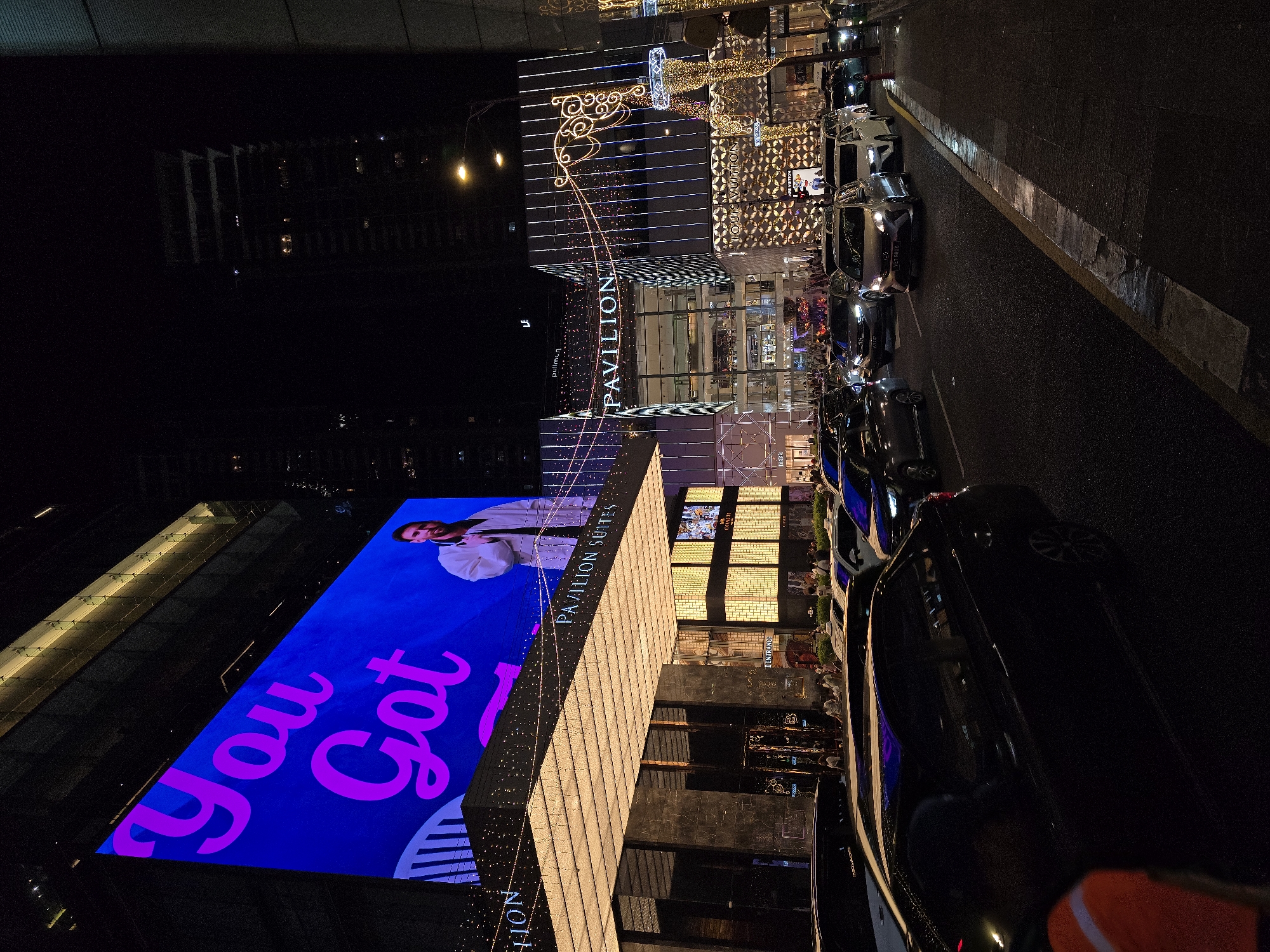 The image captures a nighttime street scene in Kuala Lumpur, Malaysia, likely in the vicinity of the Pavilion Kuala Lumpur shopping mall, given the prominent "PAVILION" signage. A large LED screen on a building displays the text "You Got" in bright pink lettering against a blue background, with a man in a white jacket also visible on the screen. Below the screen, the entrance to "PAVILION SUITES" is adorned with decorative lights. The street is filled with a traffic jam of various cars, including sedans and SUVs, illuminated by streetlights and the glow from buildings and billboards. Pedestrians are visible on the sidewalks, and the architecture of the surrounding buildings suggests a busy urban environment. The overall atmosphere is one of a vibrant, illuminated cityscape at night.