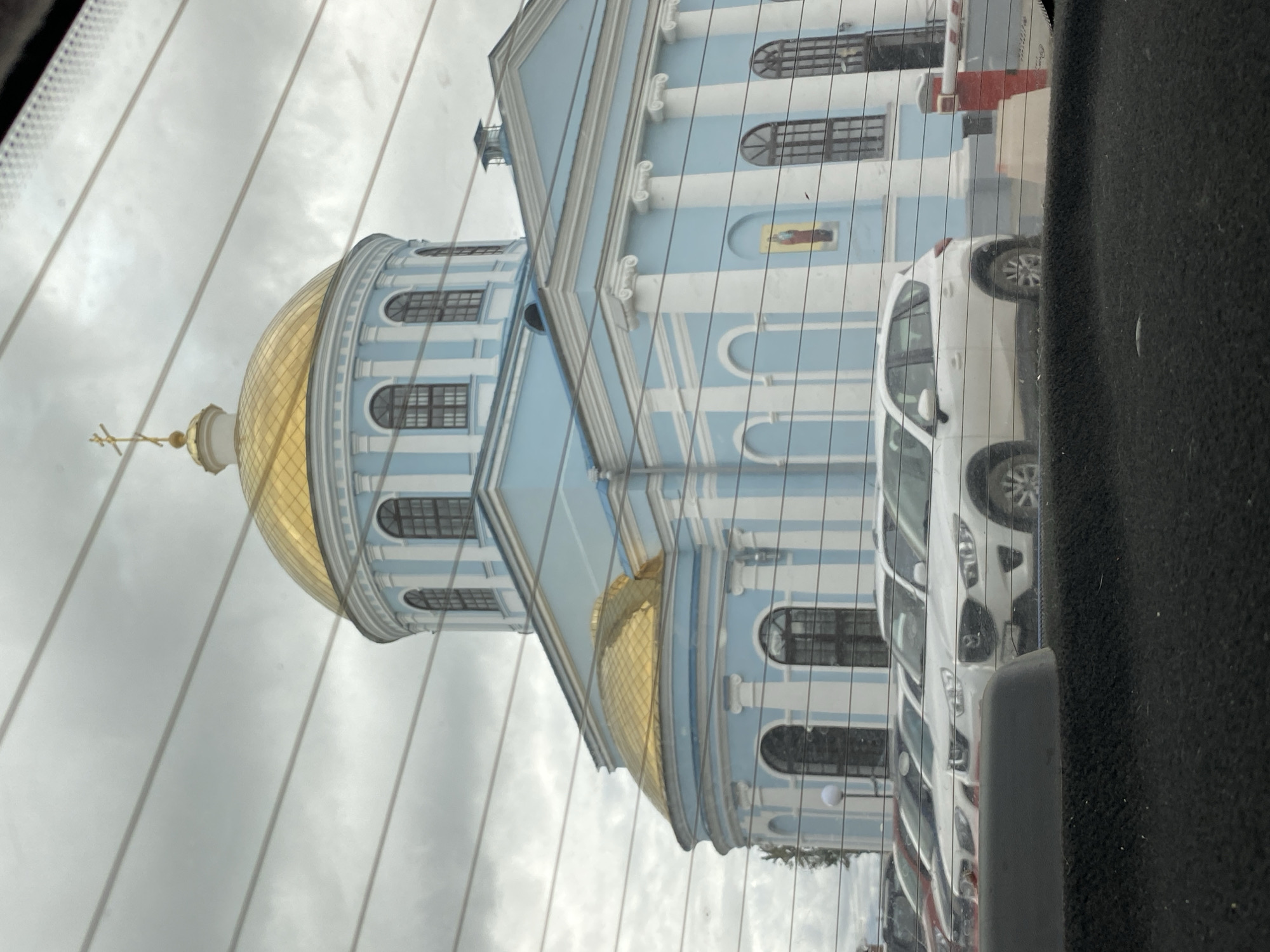 This image captured from within a vehicle shows a reflection on a car window. The primary subject is a light blue building with white columns and arched windows, topped with a golden dome. A cross is visible on top of the dome. The building's architecture suggests it is a church or cathedral.
Reflected in the window are several white cars parked in a row, and a bit of the interior of the vehicle, including a dark fuzzy material on the right side and a grey plastic edge. The image is vertically oriented, with the building's dome appearing at the top left. The sky visible through the reflection is overcast, indicating a cloudy day. The weather appears to be cool, given the attire of a person reflected in the church's window, wearing what looks like a jacket. The overall lighting suggests it is daytime.
The background of the reflection shows a parking area. The scene seems to be a view from a car parked in front of or next to a religious building, with other vehicles nearby. The reflection has a slightly distorted quality due to the glass. The image is set in Votkinsk, Russia. No visible text is present in the image.