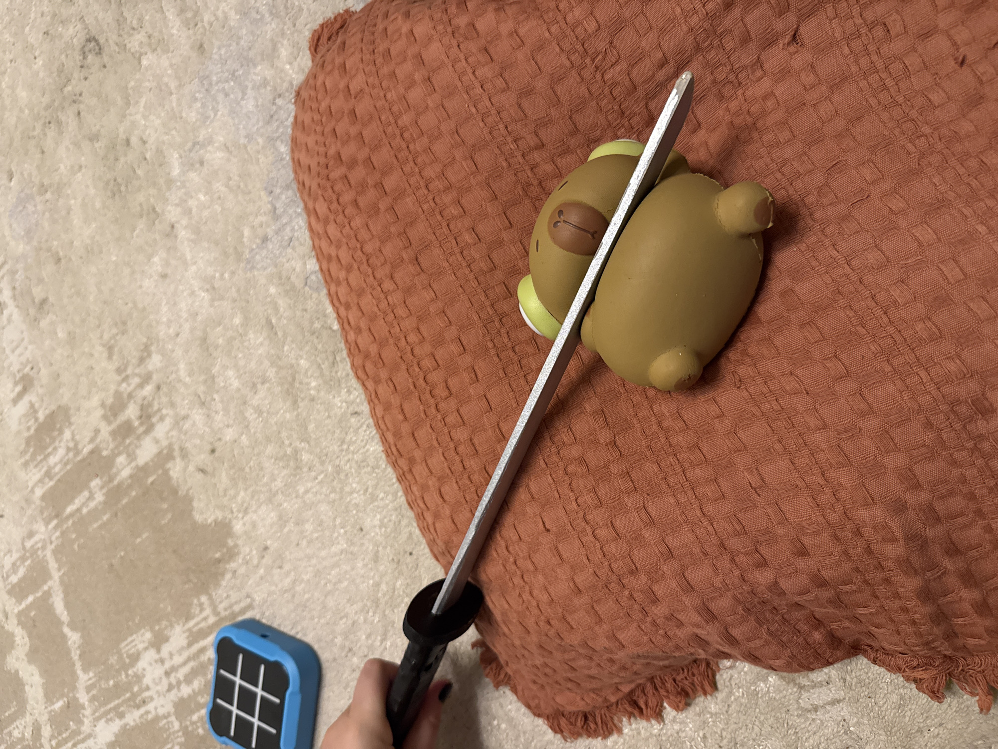 A hand holds a toy sword over a brown, squishy toy bear, positioned on a rust-colored textured fabric surface, likely a pillow or blanket, with a small blue object resembling a tic-tac-toe board in the background on a light-colored, textured floor.