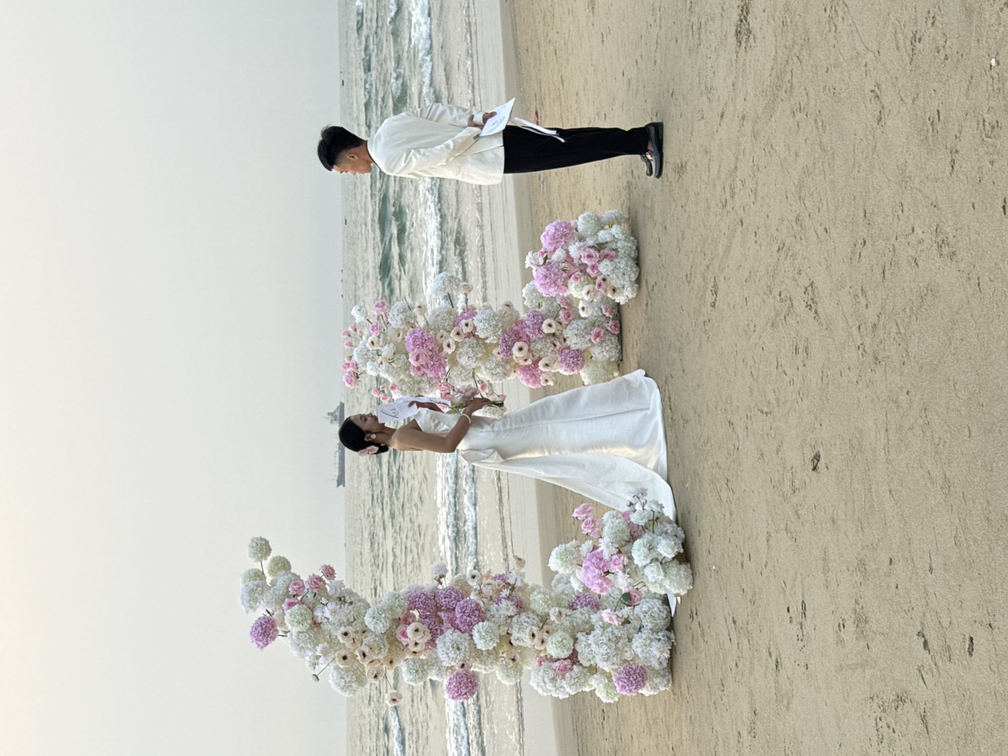 A wedding ceremony is taking place on a beach. A man, dressed in black pants, a white jacket, and black shoes, holds papers in his hands. A woman, wearing a white wedding dress, stands in front of him, holding a bouquet of flowers. They are both positioned amidst a floral arrangement of white and pink flowers. The location appears to be a beach, with the ocean visible in the background. Đà Nẵng, Vietnam, is not identifiable in the image.