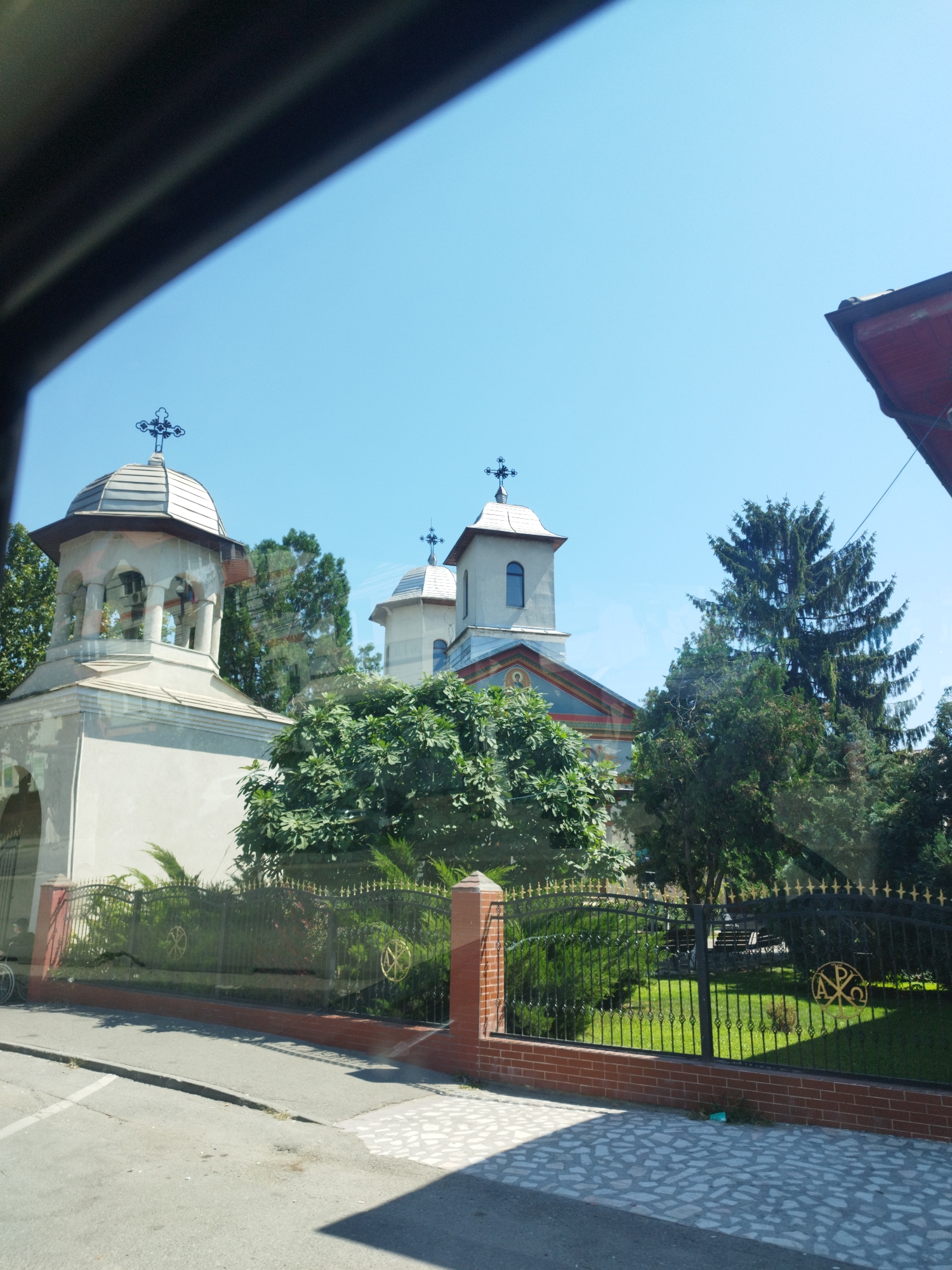 The image shows a view of a church in Bragadiru, Romania, as seen through a window. The church has multiple domes topped with crosses and features ornate architectural details. In front of the church, there is a black wrought-iron fence with gold accents and a brick base. The fence encloses a well-maintained lawn and trees. The foreground shows a paved street and sidewalk. The sky is clear and blue.