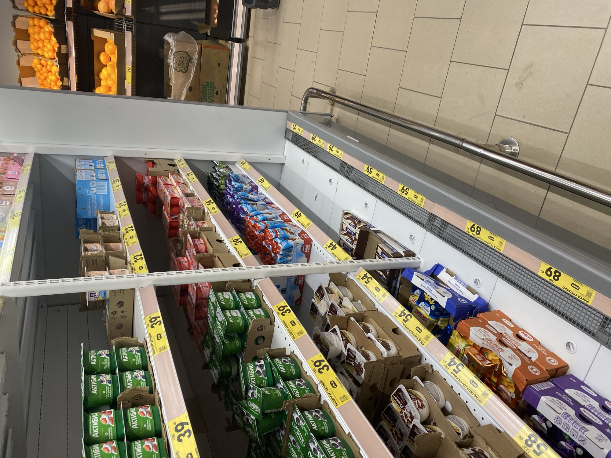 The image shows a supermarket aisle with shelves stocked with various food items, primarily yogurts and dairy products. The products are predominantly in green and white packaging, with some blue and red accents. Several price tags with yellow backgrounds are visible on the shelves, displaying prices such as "39.00", "32.00", and "54.00". On the right side of the image, shelves are stocked with oranges in cardboard boxes, with a price tag of "79.00". In the lower section, more dairy products and packaged food items are visible. The floor is tiled in a light beige color. The lighting appears to be standard fluorescent supermarket lighting. The image captures a commercial retail environment.