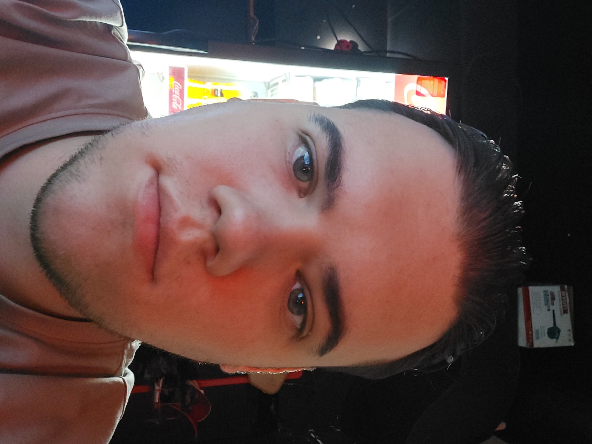 A close-up selfie of a person with light skin, dark hair slicked back, and short facial hair. The person is wearing a tan shirt. Behind the person is a Coca-Cola branded vending machine. To the right of the person, and further in the background, is a dark area containing what appears to be a small box. The picture appears to be taken indoors. There is no verifiable information in the image to confirm that the location is in Tashkent, Uzbekistan.