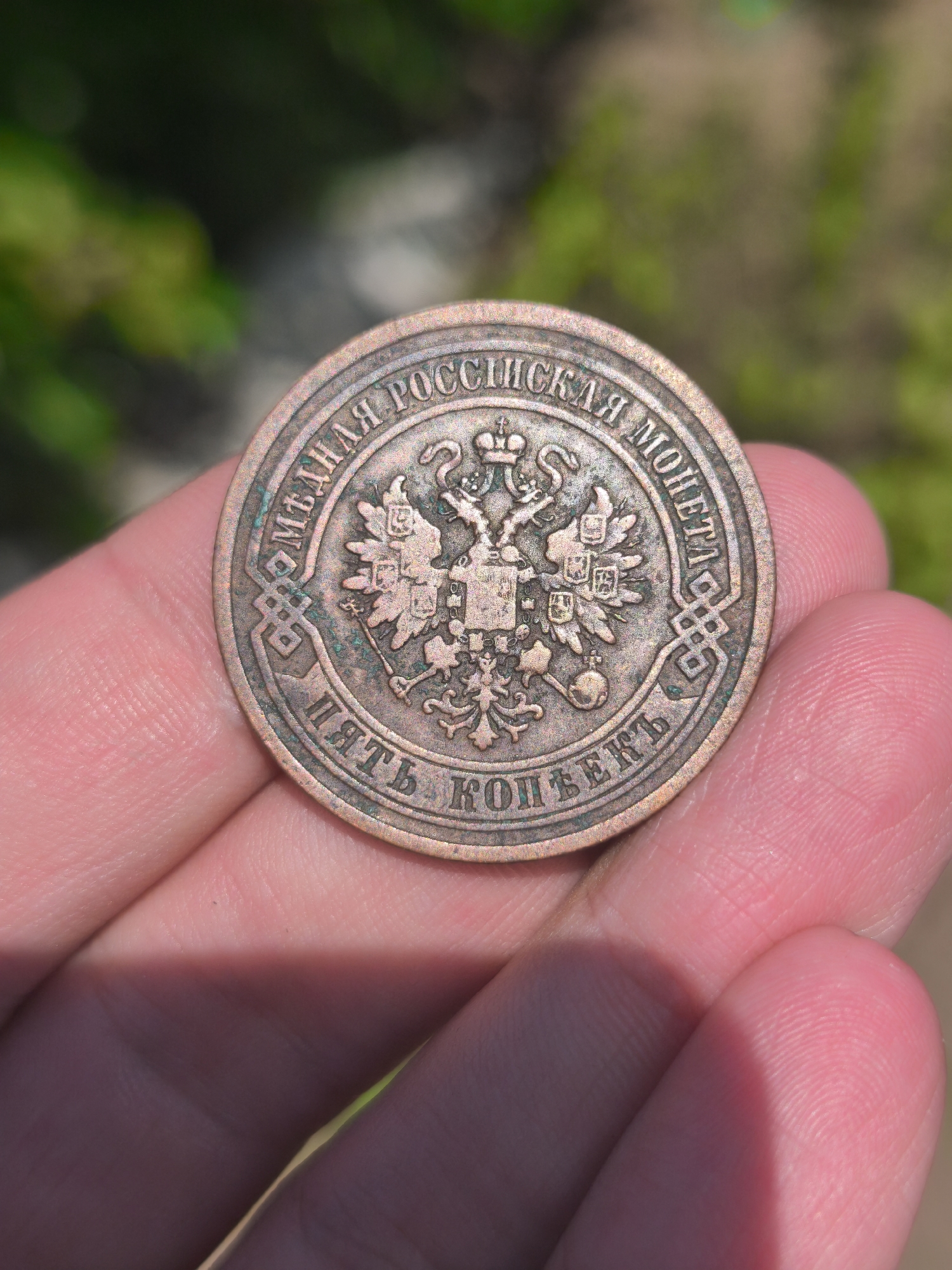 A close-up, top-down shot shows a tarnished copper coin held in a person's hand. The coin, featuring an emblem of a double-headed eagle with crowns and shields, bears Russian text that reads "РОССИЙСКАЯ МОНЕТА" (Russian Coin) and "ПЯТЬ КОПЕЕКЪ" (Five Kopecks). The background is blurred, revealing a green, out-of-focus natural environment that suggests an outdoor setting, potentially in a city like Kady, Russia.