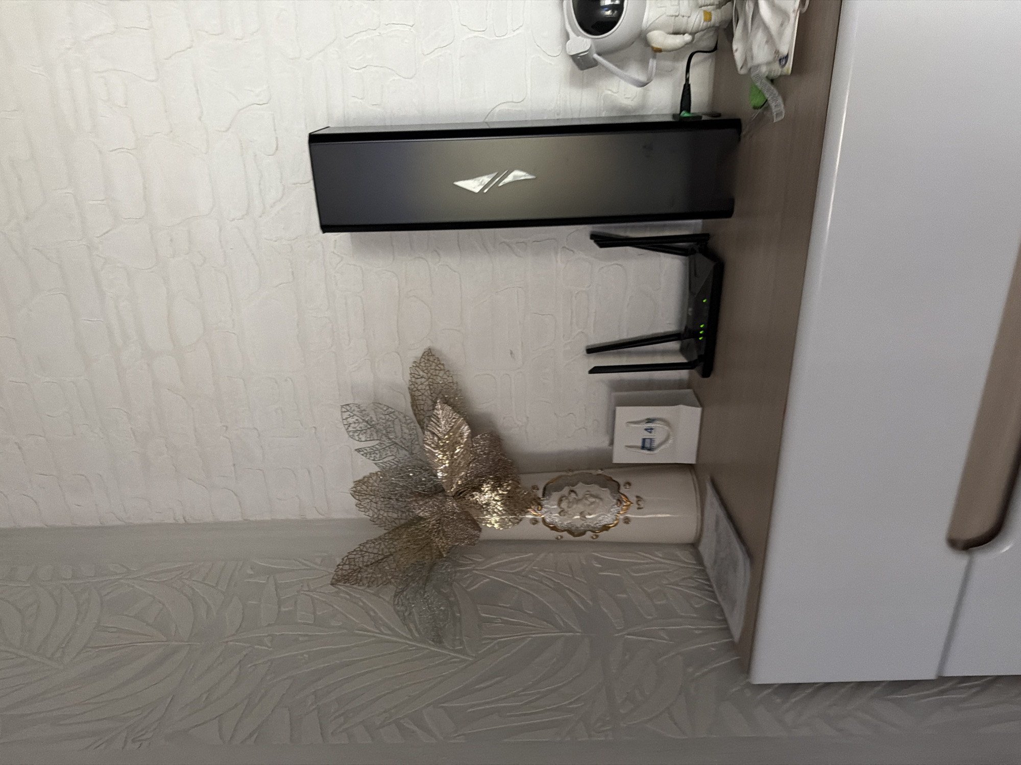 The image depicts an interior wall with several objects mounted or placed against it. The wall is painted with a textured, off-white pattern. At the top is a black box with a silver logo, mounted horizontally. Below it, there's a black Wi-Fi router with green indicator lights. Next to the router, there's a small white box. Further down, there is a white, cylindrical vase decorated with a silver bow. To the right of these objects, part of a white cabinet is visible. The location is not explicitly stated.