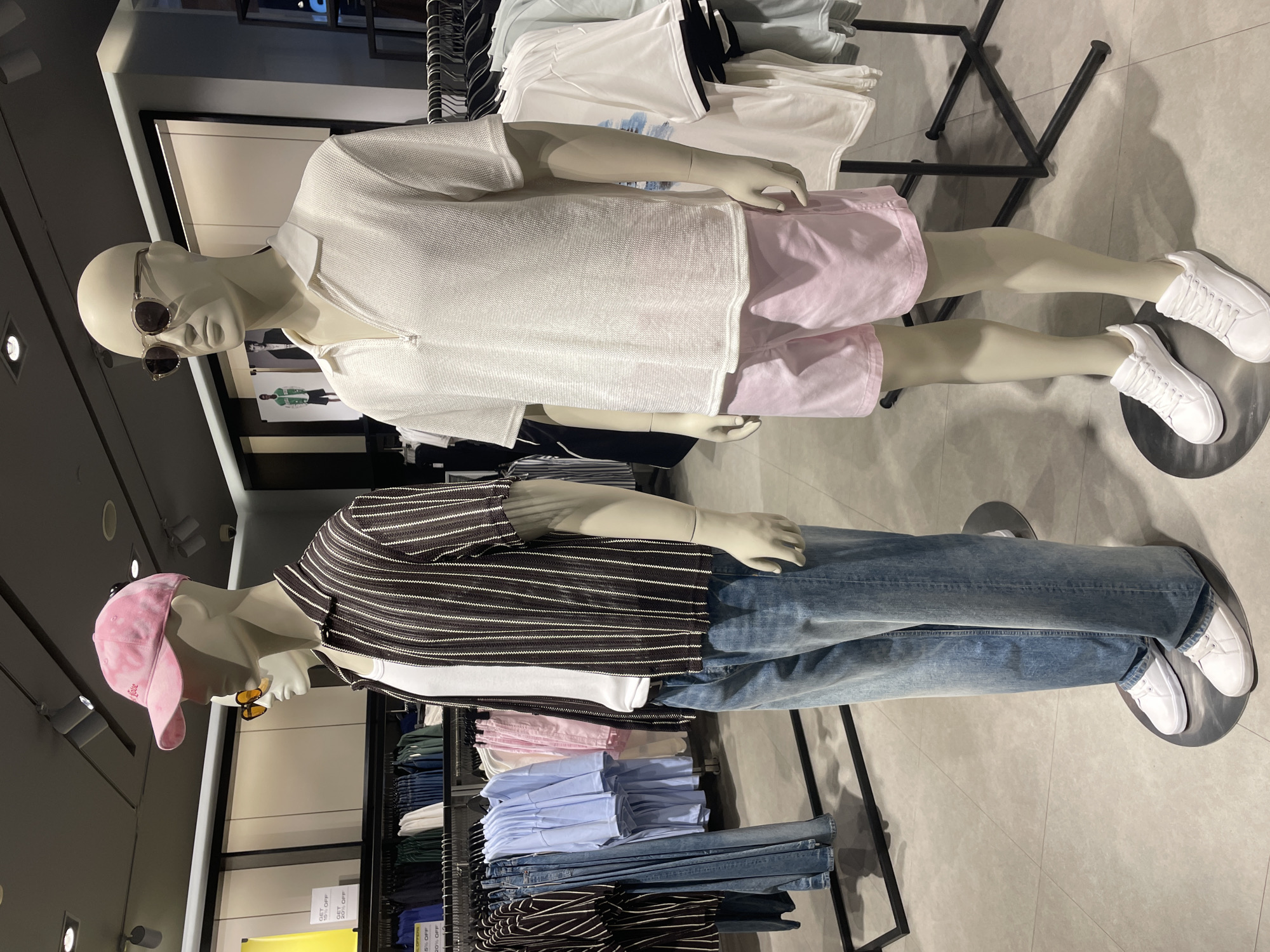 Two mannequins are prominently displayed in what appears to be a modern retail clothing store. The image is oriented vertically, showing the mannequins standing upright on a light gray tiled floor.
The first mannequin, positioned in the foreground to the right, wears a white textured polo shirt with short sleeves, light pink shorts, and white lace-up sneakers. It also sports dark sunglasses. Its arms are bent, with hands resting near its waist.
The second mannequin, slightly behind and to the left, is dressed in a dark brown or black horizontally striped short-sleeved shirt with thin white stripes, light blue denim jeans, and white lace-up sneakers. This mannequin wears a pink baseball cap with partially obscured text that appears to say "Love..." and small yellow-tinted sunglasses. Its arms are also bent, with hands near its waist.
In the background, behind both mannequins, are metal clothing racks holding folded garments, including stacks of white, light blue, and pink shirts, as well as blue jeans. The store features a clean aesthetic with white walls and exposed black metal fixtures. Artificial lighting illuminates the scene from overhead.
Visible text includes "GET 15% OFF" repeated multiple times on a yellow sign in the background, and partial text on the pink baseball cap that reads "Love...". The overall scene depicts a retail clothing display showcasing casual fashion items.