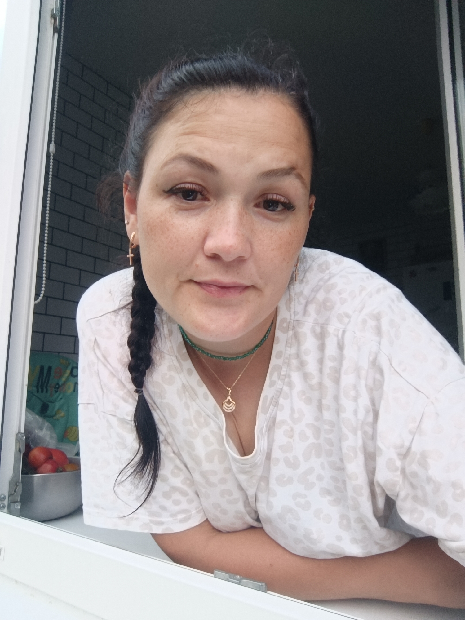 The image shows a woman with dark hair in a braid, wearing a white t-shirt with a leopard print pattern. She has freckles on her face and is wearing gold cross earrings and a necklace with a gold pendant. She is leaning on a window frame. In the background, there is a tiled wall and a bowl of tomatoes on a counter. The image appears to have been taken outdoors, possibly in a residential area of Chaikovsky, Russia. The lighting suggests it is daytime.