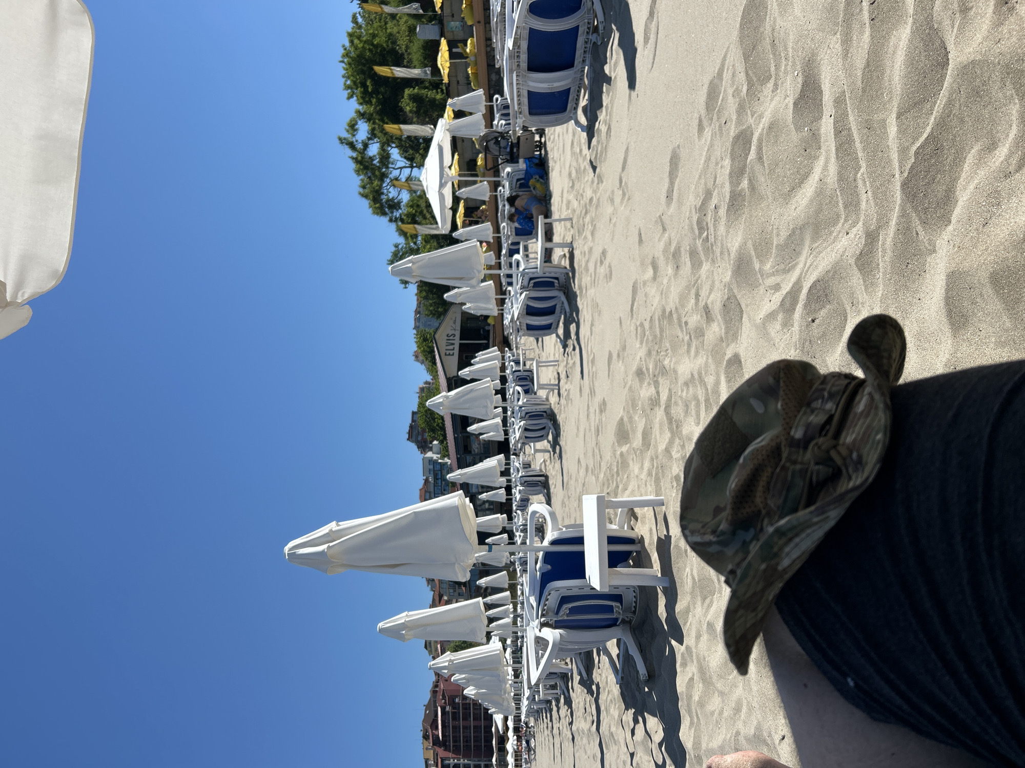 This is a low-angle, eye-level shot of a sandy beach in Sozopol, Bulgaria, on a clear, sunny day. In the foreground, a person's leg and a camouflage-patterned bucket hat are visible. The beach is lined with rows of white beach umbrellas and blue and white lounge chairs, many of which are folded and stacked. In the middle ground, a few people are visible near a beach cafe with the sign "ELVIS" on it. Beyond the cafe, trees and buildings can be seen in the distance, suggesting an urban or resort area. The sand is light brown and appears dry and soft, with visible footprints and shadows cast by the umbrellas and chairs. The sky is a bright, cloudless blue.