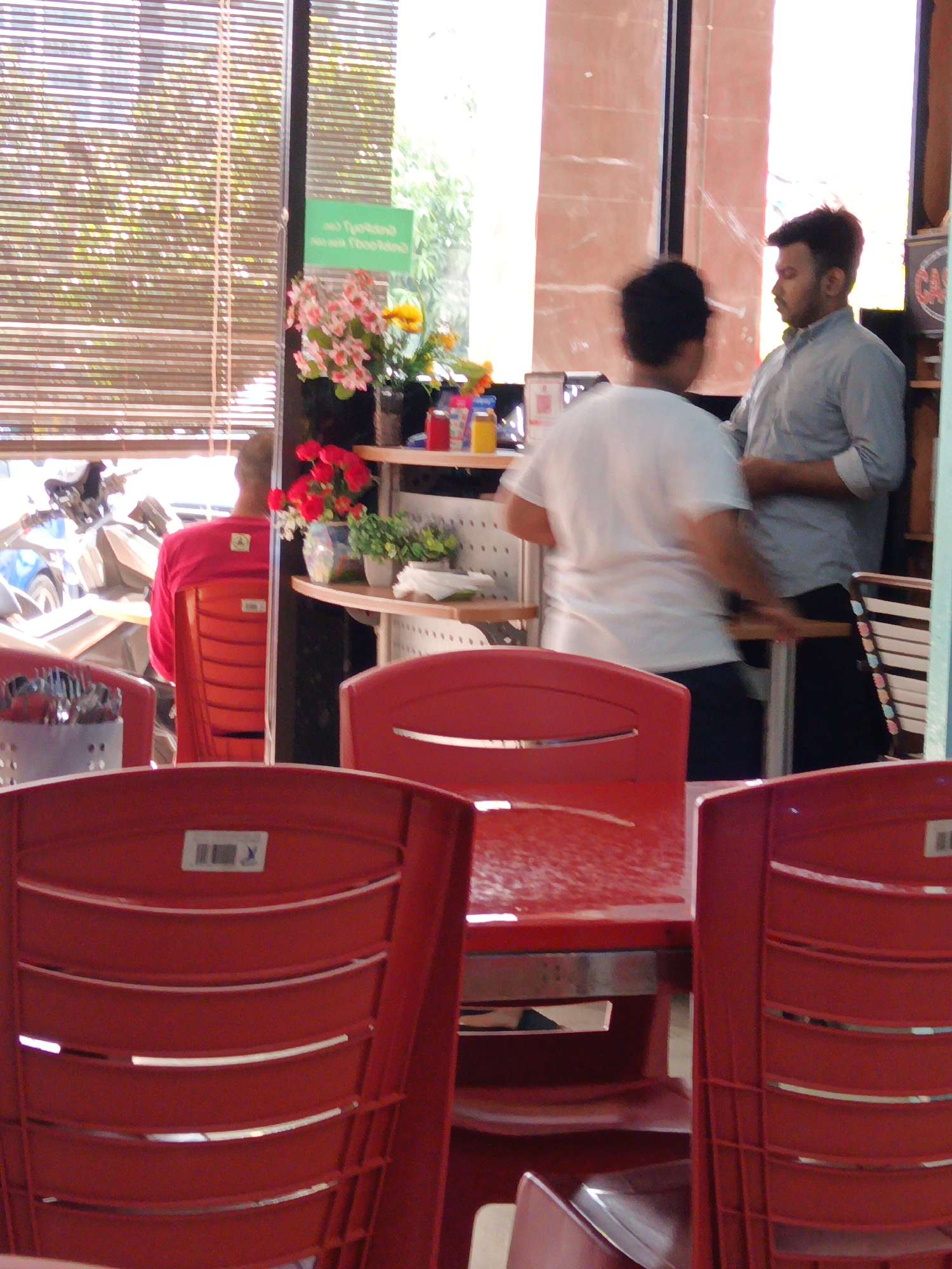 The image shows the interior of a cafe or restaurant with red plastic chairs and tables. In the background, two men are standing. One man, wearing a light blue shirt, is looking towards the right, while the other man, wearing a white t-shirt, is facing away from the camera. Behind them, there is a window with blinds, and a shelf with flowers and bottles. The scene suggests a casual indoor setting, possibly in Kuala Lumpur, Malaysia, given the context of the cafe culture in the city.