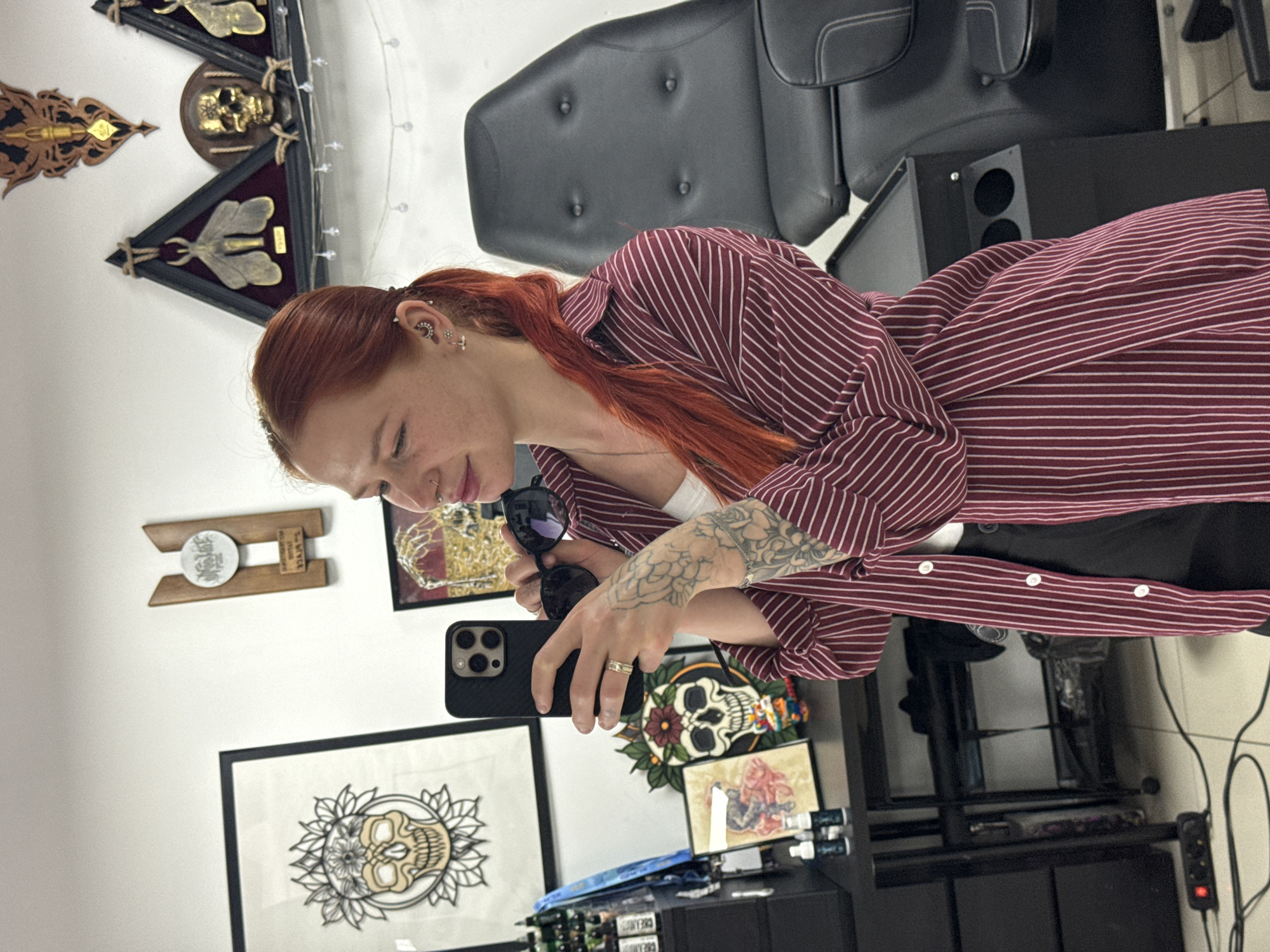 A woman with auburn hair is standing indoors in what appears to be a tattoo parlor. She is wearing a maroon and white striped button-down shirt and is holding a cell phone in her right hand, and a pair of black sunglasses in her left. Her left arm is covered in tattoos.
Several framed pieces of artwork adorn the walls behind her, including drawings of skulls and various other designs. Other decorative items are present on the walls. A black tattoo chair is visible in the background.
The location is possibly in Rostov-on-Don, Russia.