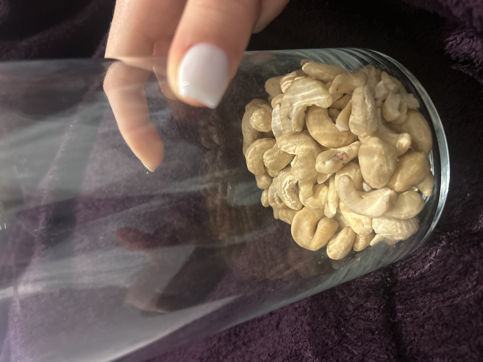 A close-up shot shows a glass container partially filled with cashew nuts. A person's hand is holding the container. The background appears to be a purple textured material. The location and city are not discernible from the image.