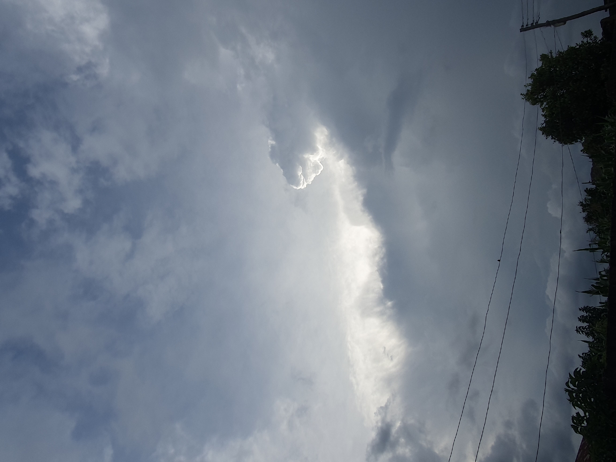 This is a photograph of a cloudy sky over Gura Top, Nigeria. The clouds are dark and heavy, suggesting an impending storm. A patch of bright light shines through the clouds in the upper right quadrant of the image. In the foreground, power lines stretch across the frame, with a small bird perched on one of them. Below the power lines, silhouetted trees and vegetation are visible. The overall impression is one of dramatic weather, with a sense of anticipation.