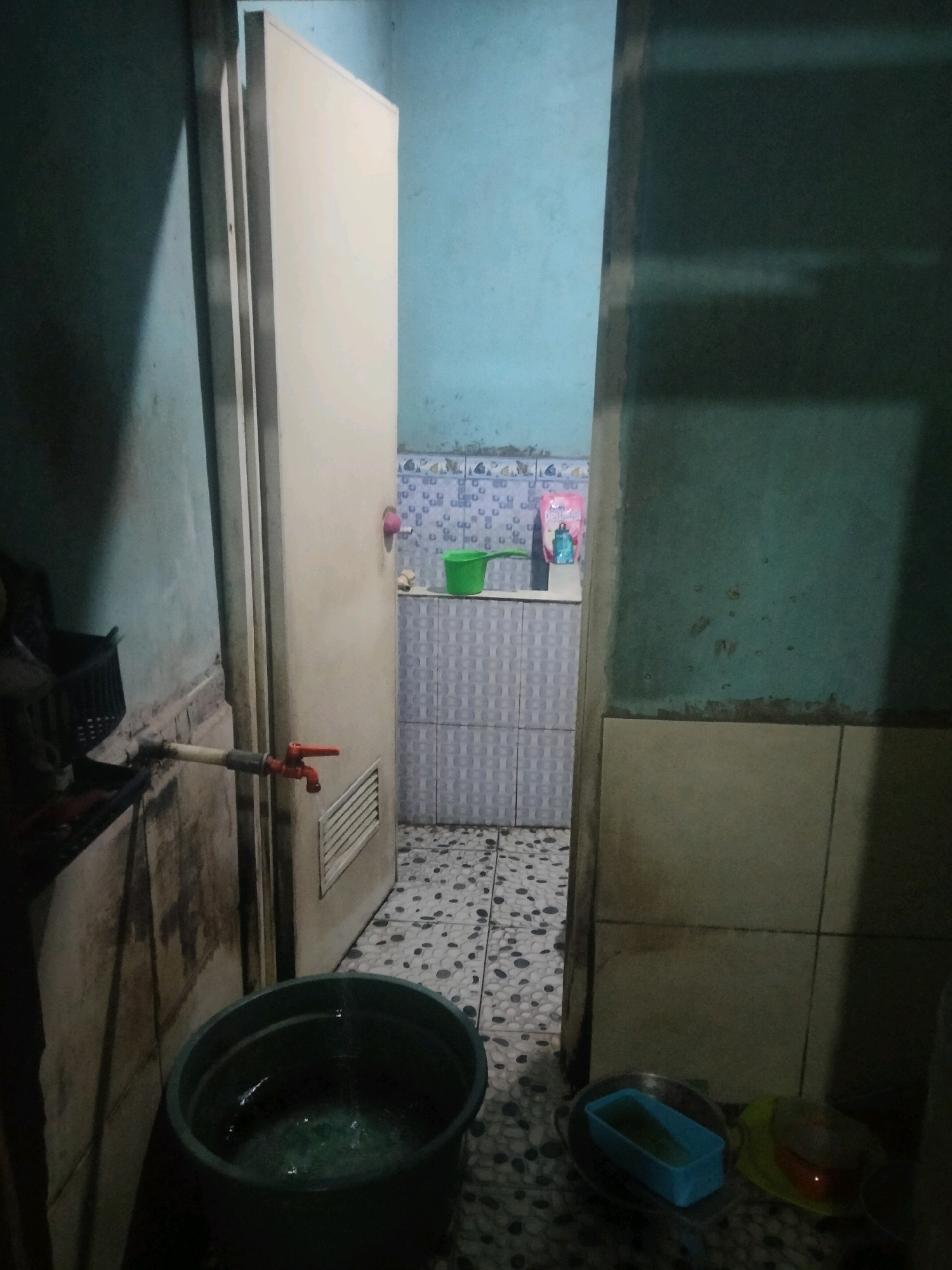 In a dimly lit room in Suka Sari, Indonesia, a water faucet is attached to a pipe that extends horizontally from a wall and into the frame. The faucet is red and appears to be made of plastic. Below the faucet, a large dark green plastic tub is positioned, collecting dripping water. The floor is covered in small, round, grey and white tiles, with a mosaic-like pattern. To the right of the tub and faucet, a door is ajar, revealing a small, tiled bathroom. The bathroom walls are a light blue, with a band of patterned tiles near the ceiling. A green bucket sits on a tiled surface inside the bathroom, next to a pink and blue package, possibly of cleaning supplies. The overall environment suggests a functional, albeit somewhat cluttered, living space, likely a kitchen or utility area adjacent to a bathroom.
