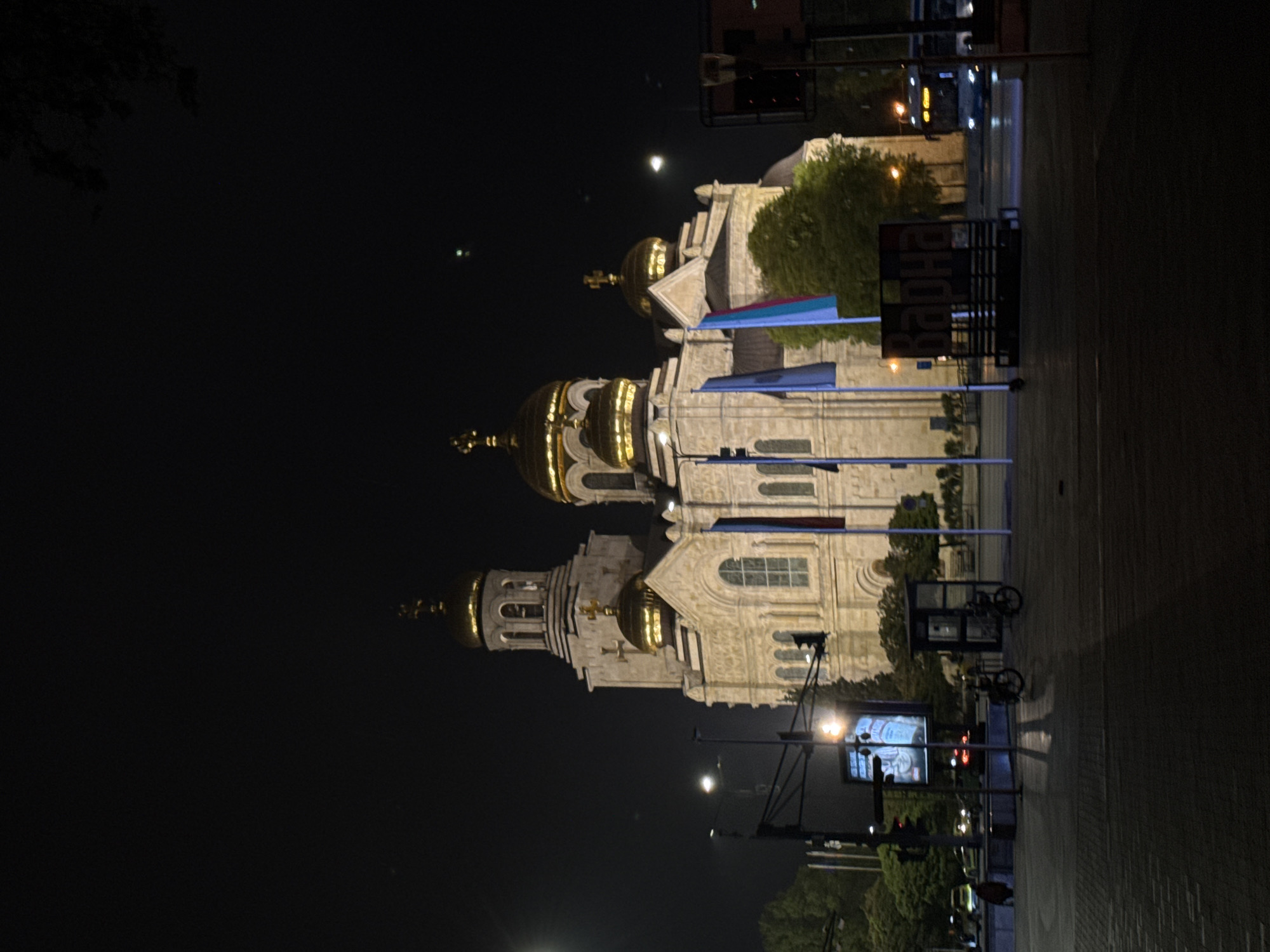 The image shows the Cathedral of the Assumption of the Virgin in Varna, Bulgaria, at night. The cathedral's golden domes are illuminated against the dark sky. Blue and white flags are prominently displayed in front of the building. To the right, a paved area is visible, along with a structure that appears to be a bus stop or similar urban installation. There are trees, streetlights, and an electronic advertising display in the background. The scene is dark and primarily lit by the cathedral itself, streetlights, and the electronic display.