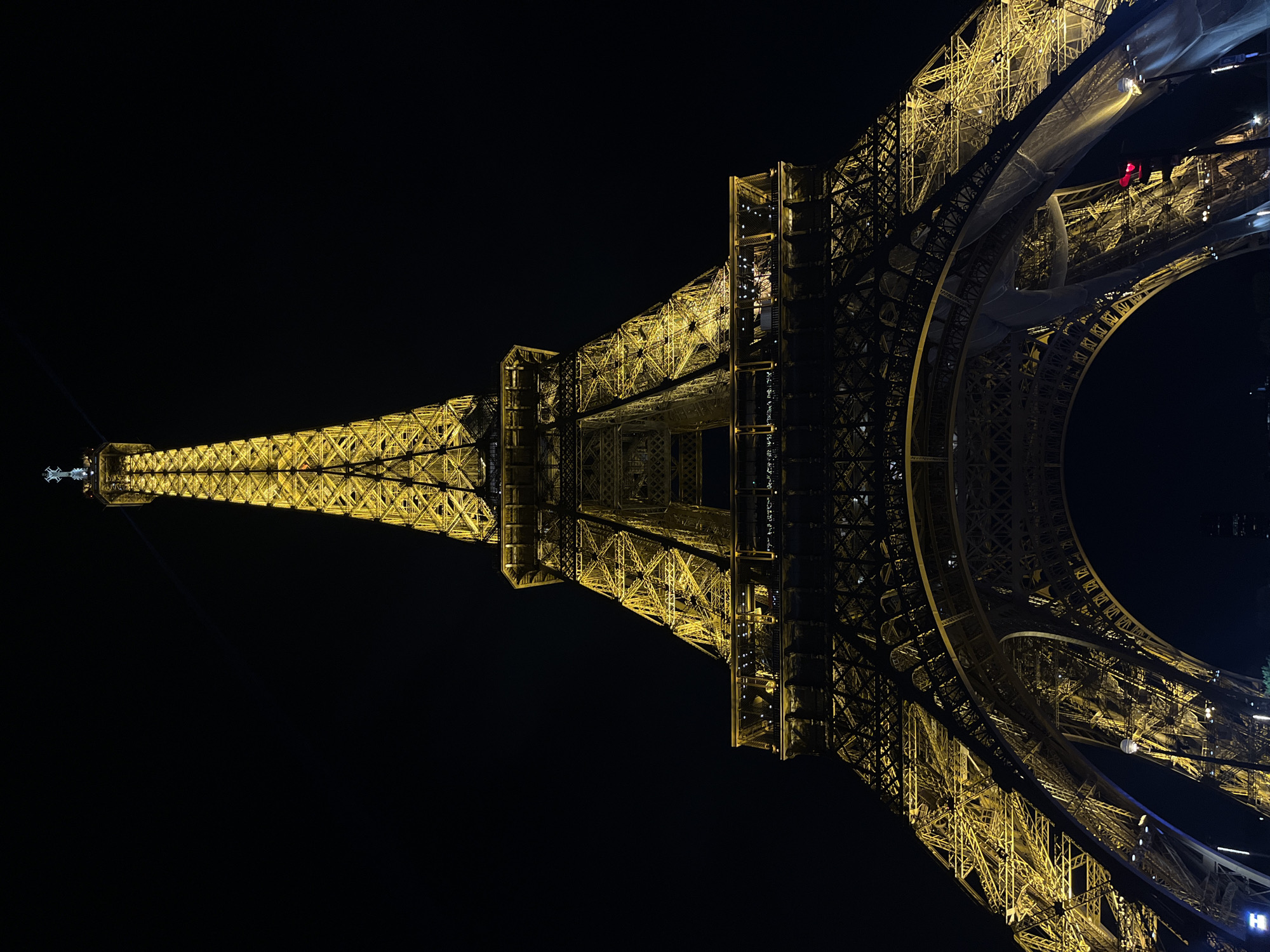 The image captures the iconic Eiffel Tower in Paris, France, at night. The tower stands tall against a completely dark, clear sky, illuminated entirely by a warm, golden light that accentuates its intricate wrought-iron lattice structure. A bright beam of light emanates from the very top of the tower, projecting upwards and outwards like a beacon.
The various crops offer closer views of the tower's detailed construction. The geometric patterns of the ironwork are clearly visible across different sections, with varying levels of brightness highlighting the depth and complexity of the architecture. Some parts of the tower, particularly in the mid-to-lower sections, show translucent white netting or scaffolding draped over the framework, suggesting ongoing maintenance or construction work. A solitary red light, potentially a navigation light or related to the temporary structures, is visible in one of the lower arches. No people or discernible text are present in the image. The scene conveys the grandeur and structural marvel of the landmark under a clear night sky.