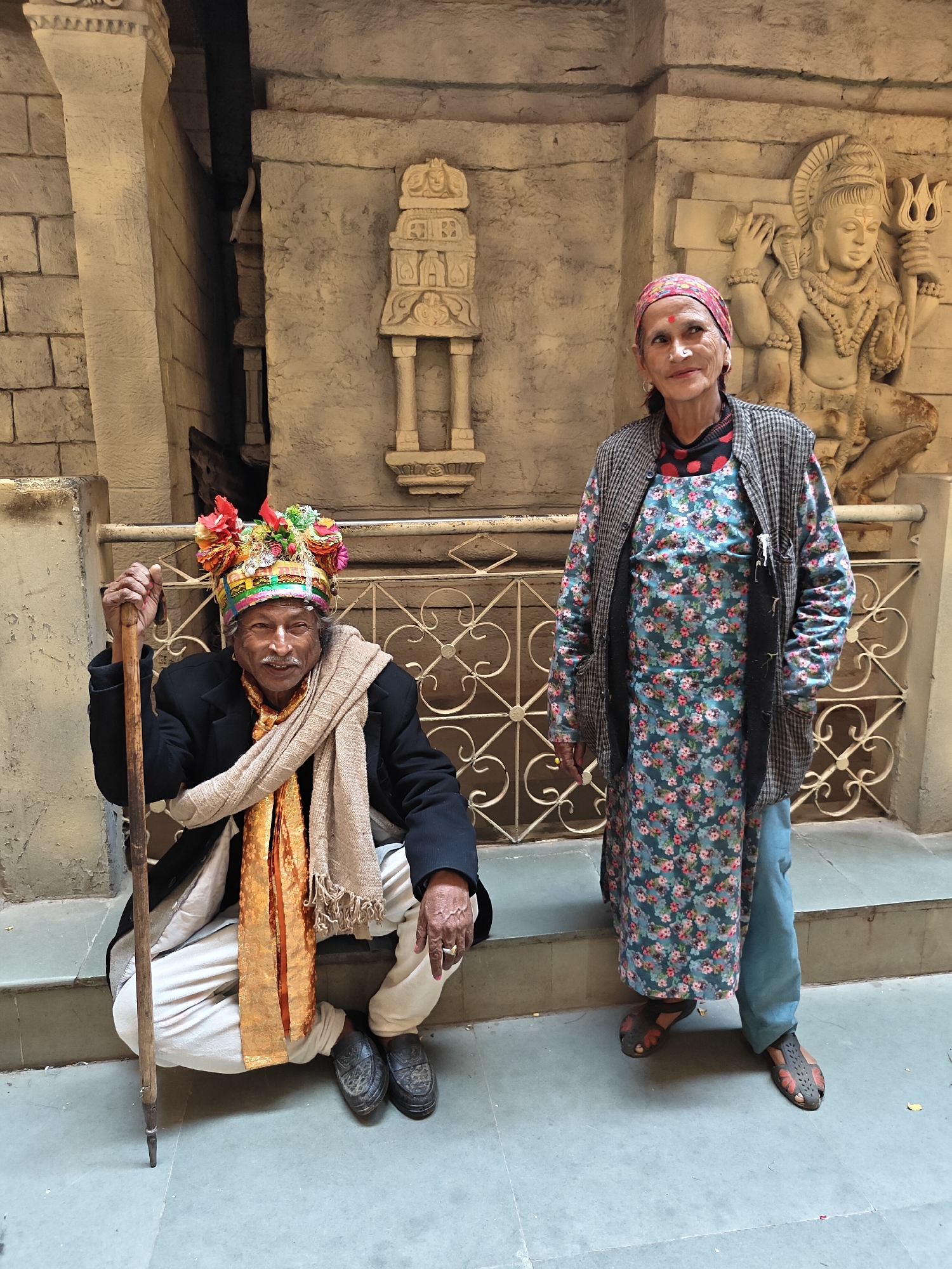 An elderly man and woman are posing for a photograph in what appears to be a stone temple complex, likely in Manikaran, India, as suggested by the location context.
The man is positioned on the left, squatting on a stone step. He holds a tall, slender wooden staff in his right hand. He is wearing a distinctive, colorful headpiece adorned with vibrant floral decorations, a black jacket, a light beige shawl draped around his neck and shoulders, and an orange patterned scarf underneath. He has a mustache and a bindi-like mark on his forehead, and he looks towards the camera with a gentle smile. His footwear consists of dark, possibly leather, slip-on shoes.
Standing to the right of the man is an elderly woman. She wears a patterned red and white headscarf, a long-sleeved blue tunic with a prominent floral pattern, light blue pants, and a grey and black checkered open jacket. She has a nose ring and a red bindi on her forehead. Her footwear consists of open-toed sandals.
The background is dominated by aged stone architecture. To the man's left, a narrow, dark passageway is visible between high stone walls. Behind the woman, there is an intricate stone carving of a deity, resembling Shiva, depicted with a trident in one hand and a cobra around his neck. A decorative metal railing separates the foreground from the back wall. The ground is a light-colored tiled surface, with stone steps behind the man. The lighting indicates it is daytime, possibly under an overcast sky or in a shaded area. No visible text is present in the image.