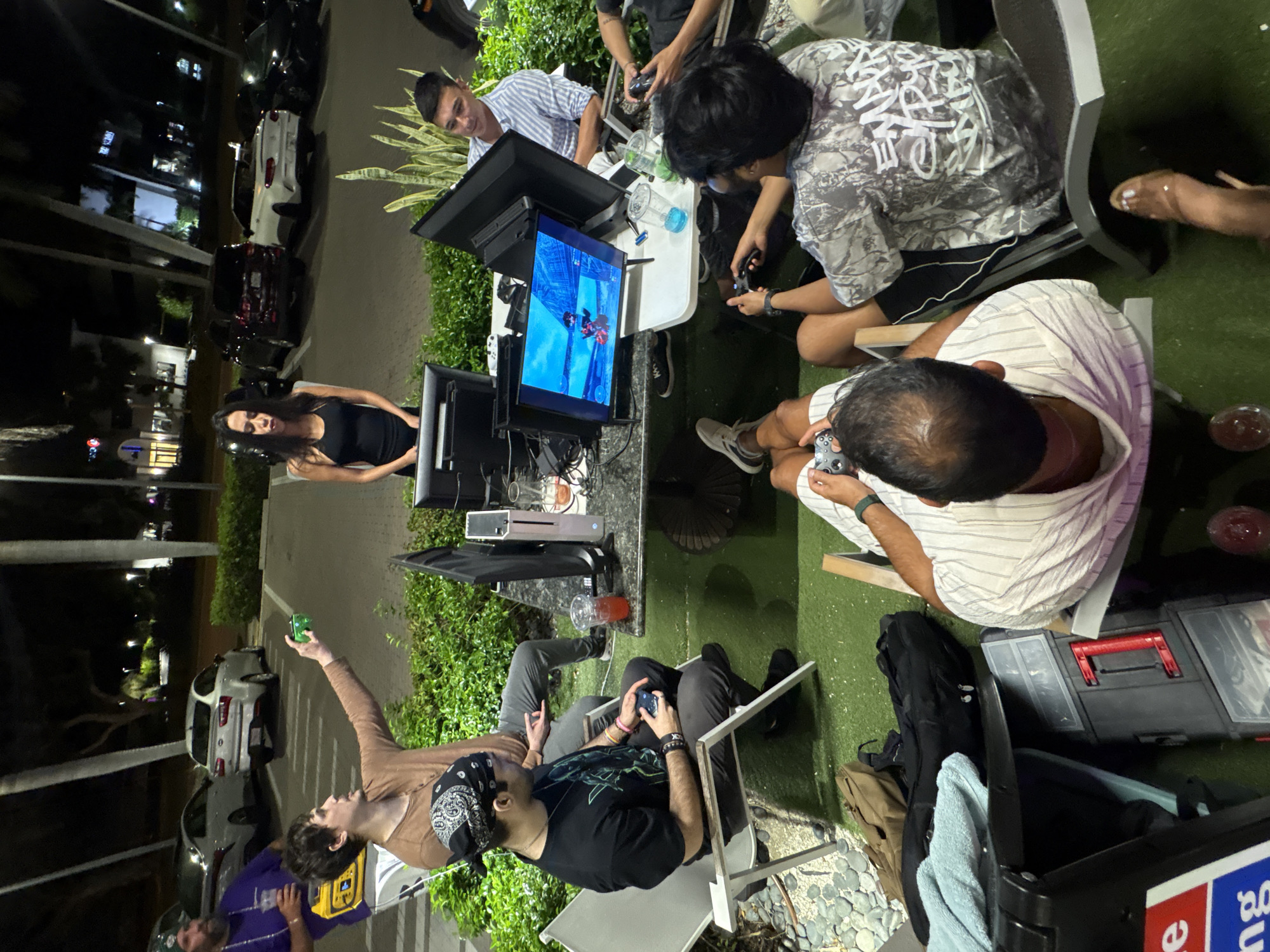 The image depicts an outdoor video gaming session taking place at night. Several individuals, primarily young adults, are gathered around multiple screens, engaged in playing or observing.
In the mid-ground, two men are intently focused on a monitor displaying a colorful video game, possibly an action or fighting game with animated characters. One, wearing a striped shirt, sits facing the screen, while another, in a camouflage-patterned shirt, leans in, both holding game controllers. Another man, seen from an overhead perspective, in a white striped shirt, is also holding a controller, suggesting active participation.
To the left, a woman in a black tank top stands and looks towards the left side of the frame, appearing to be in conversation. Further to the bottom-left, a man in a brown long-sleeve shirt holds up a small green object. Beside him, another man in a black T-shirt and a bandana holds a controller. A person in a purple shirt is partially visible in the bottom-left corner.
The setting is an outdoor area, likely a patio or paved space, covered in artificial green turf and paving stones. The time is clearly night, as evidenced by the darkness in the background and the reliance on artificial lights illuminating the scene. Several cars are parked along what appears to be a street in the background.
Equipment visible includes multiple gaming monitors/TVs, controllers, tables, and chairs. Drinks in clear cups, one with blue liquid and another with red, are on the tables. A large black storage box with red latches is visible on the ground, along with a smaller black case that has partial text "BU" and "e" visible on a label. A yellow power bank and various cables complete the setup. One person's shirt has stylized text that might read "WOKE." The overall mood appears casual and social, centered around shared gaming activity.