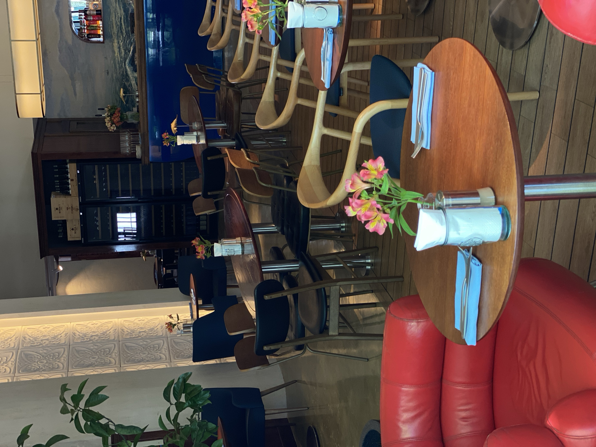 This image captures the interior of a restaurant or bar in Sochi, Russia, appearing to be set up for service during the daytime. No people are visible.
The scene features numerous round, polished wooden tables with tall metal bases, each set with light blue napkins, silverware, and a small vase holding fresh pink and yellow flowers, possibly alstroemeria. Seating includes a variety of chairs: some with light wooden, curved frames and dark upholstered seats, and others with more traditional wooden designs. A prominent seating area in the foreground consists of plush red leather armchairs or a sofa, contributing a vibrant color accent.
Along the back wall, a long, deep blue bar counter is visible, above which a mural depicting a tranquil seascape or ocean scene stretches across. Below the counter, a dark wooden cabinet houses professional wine coolers filled with bottles; legible text on a wooden box inside reads "RODAI" and "CABERNET." The floor throughout the establishment is covered in light-colored wooden planks, reminiscent of a ship's deck. A large, rectangular light fixture illuminates the space from above. A lower section of a side wall features decorative white embossed tiles with a repeating, intricate pattern. Green potted plants are strategically placed, adding natural elements to the inviting ambiance. The establishment appears well-maintained and ready to welcome guests.
