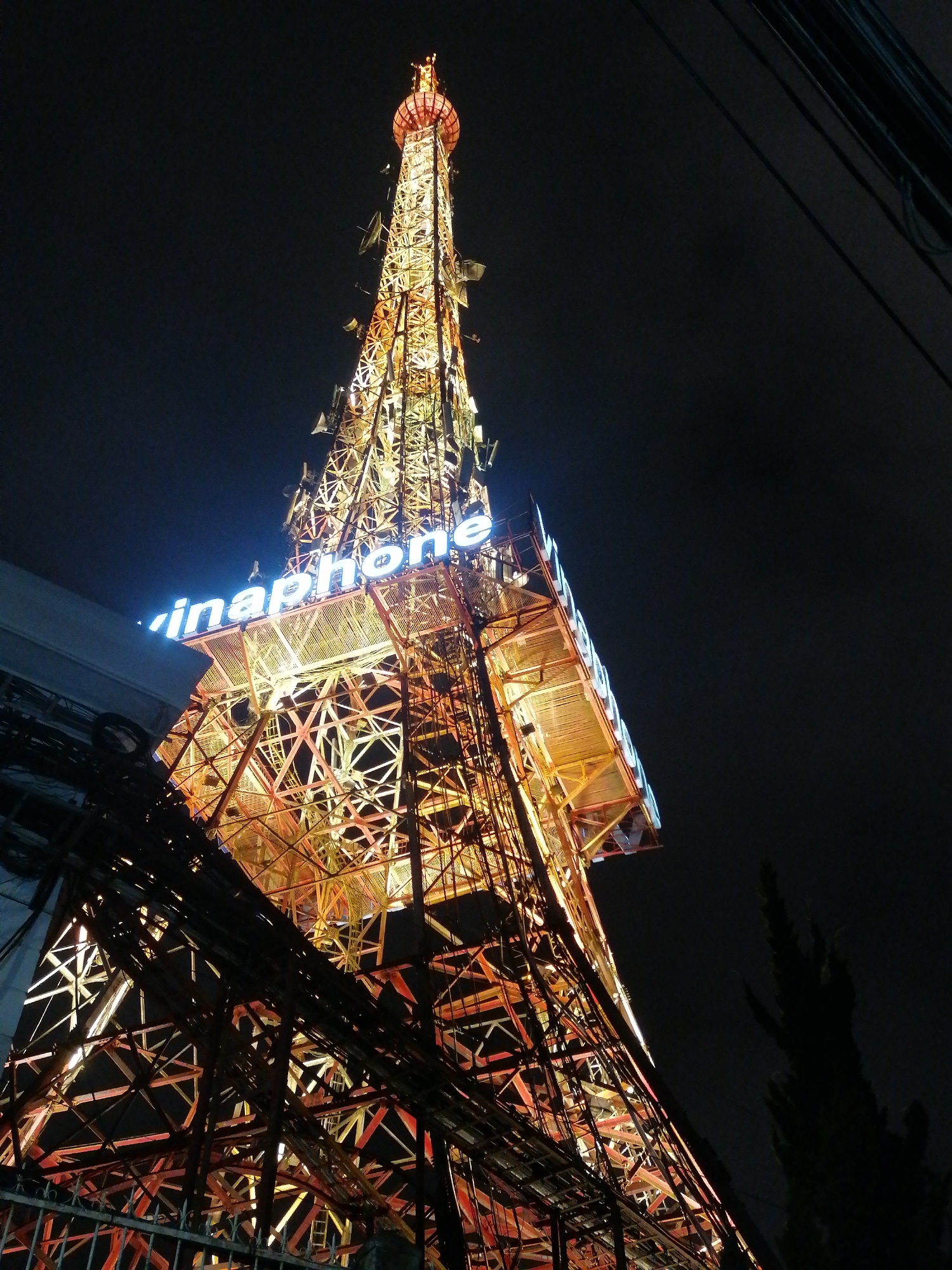 This image captures a towering telecommunications mast, brightly illuminated against a dark night sky, in Da Lat, Vietnam. The primary subject is a large, intricate lattice-work tower, constructed from metal beams, which glows with a warm, yellowish-orange light from within, emphasizing its complex structure.
Numerous satellite dishes and various antennas are affixed along the height of the tower, particularly visible in its upper sections, indicating its function for communication. Around the mid-section, the word "Vinaphone" is prominently displayed in vibrant blue neon letters, identifying the Vietnamese mobile network operator.
The perspective is looking upwards from a low vantage point, enhancing the tower's imposing scale. The time of day is clearly nighttime, evidenced by the deep black sky and the reliance on artificial illumination. In the foreground, a dense tangle of dark electrical cables and wires stretches across the frame, with some seemingly connected to a dark building or structure partially visible on the far left. At the very bottom, a dark metal fence runs horizontally, separating the viewer from the base of the tower. To the far right, the dark silhouette of a tall, slender tree stands against the night sky. The scene is static, showcasing the illuminated urban infrastructure.