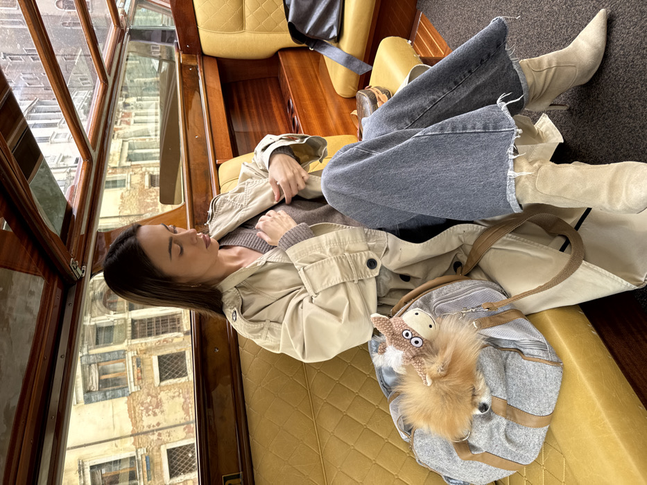 A female subject is seated inside a water taxi in Venice, Italy. She is positioned facing left, with her head inclined downwards, appearing pensive or focused on her hands. She wears a light beige trench coat over a V-neck brown sweater, dark grey wide-leg jeans with frayed hems, and light beige pointed-toe high-heeled ankle boots. Her hands are resting on or adjusting the front of her trench coat. To her left, on the yellow quilted seat, a grey and brown striped pet carrier bag contains a small, fluffy, reddish-brown dog, with only its head visible. A knitted donkey-shaped charm with white eyes is attached to the carrier bag. A dark brown handbag is partially visible on the seat to her right. The boat's interior features polished wooden paneling, yellow upholstered seating, and a dark carpeted floor. Through the boat's windows, typical Venetian architecture is visible, consisting of multi-story buildings with aged, light-colored facades and numerous windows, suggesting a canal-side view.