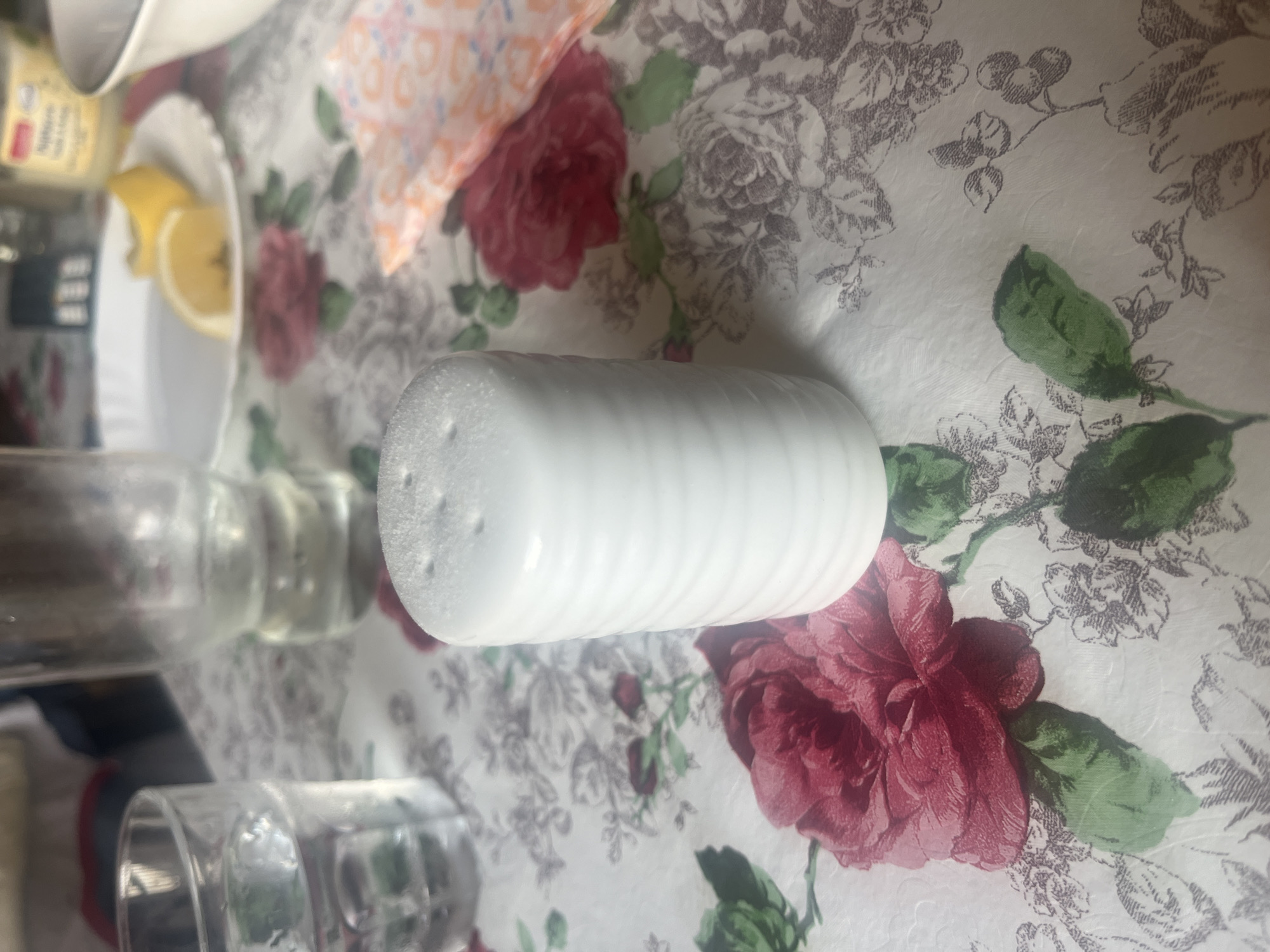 A white, ribbed, cylindrical salt shaker with visible salt crystals on its perforated top is centrally positioned on a patterned tablecloth in an indoor setting. The tablecloth features a design of large deep red roses with green leaves, interspersed with smaller, intricate grey-purple floral and leafy outlines on a white background. To the left of the salt shaker, a clear glass partially filled with water is visible, alongside a taller, out-of-focus clear glass or bottle further back. In the upper right background, a white plate holds two slices of lemon. Behind the plate, a blurred yellow container with some visible text and a dark, rectangular object are present. A portion of a white bowl is visible on the far right. To the immediate right of the salt shaker, a folded napkin with an orange and white patterned design is observed. The scene is well-lit, located in the city of Burgas, Bulgaria.