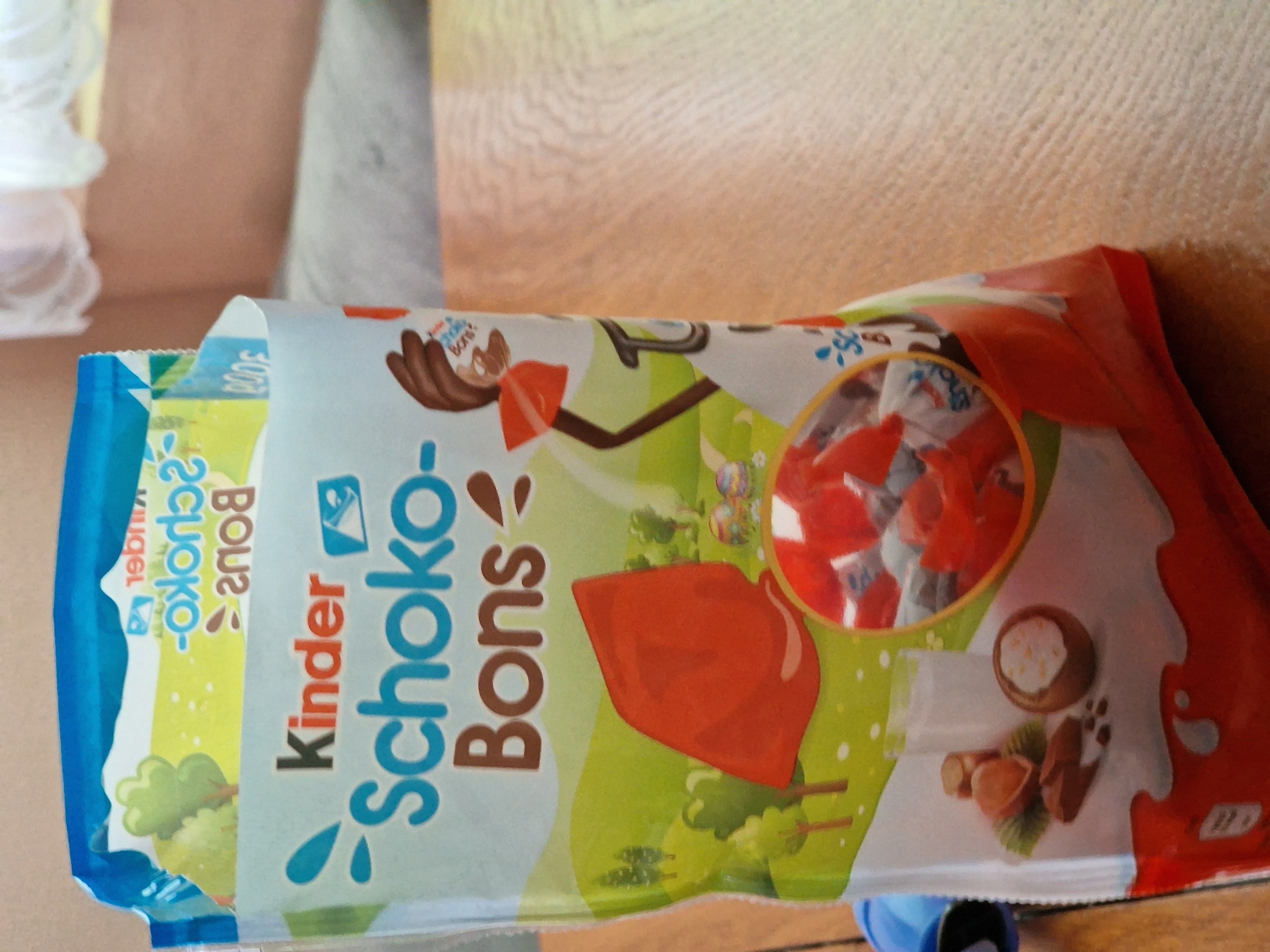 A horizontal medium shot depicts a bag of Kinder Schoko-Bons candy resting on a surface. The packaging is brightly colored with a combination of blue, green, white, and orange hues. "Kinder Schoko-Bons" is prominently displayed on the bag in large, stylized lettering. Graphics on the packaging include images of trees, hazelnuts, and the candy itself. The bag appears to be slightly open, revealing additional candy inside. The background is blurred, showing a tan-colored surface.
