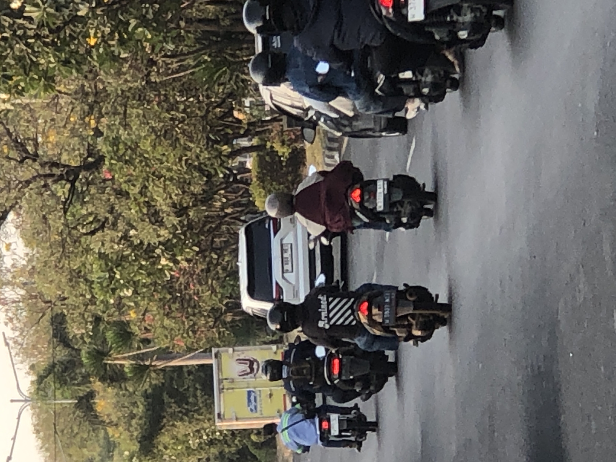 The image depicts a street scene with multiple motorcycles and cars in motion, likely in an urban environment in Ngaban, Indonesia. In the foreground, several individuals are riding motorcycles. The first motorcycle on the left has a rider wearing a blue jacket and a helmet. To their right, another motorcycle carries a rider with a black jacket and helmet, and a passenger with a helmet. Further to the right, a motorcycle is ridden by a person in a dark jacket and helmet, and behind them, another motorcycle with a rider wearing a dark jacket and helmet is visible. The road surface appears to be asphalt. In the background, a white SUV and a yellow truck are also present. The environment is lined with trees and greenery, suggesting a location with vegetation. The lighting indicates it is daytime, possibly late afternoon, given the warm tone of the light.