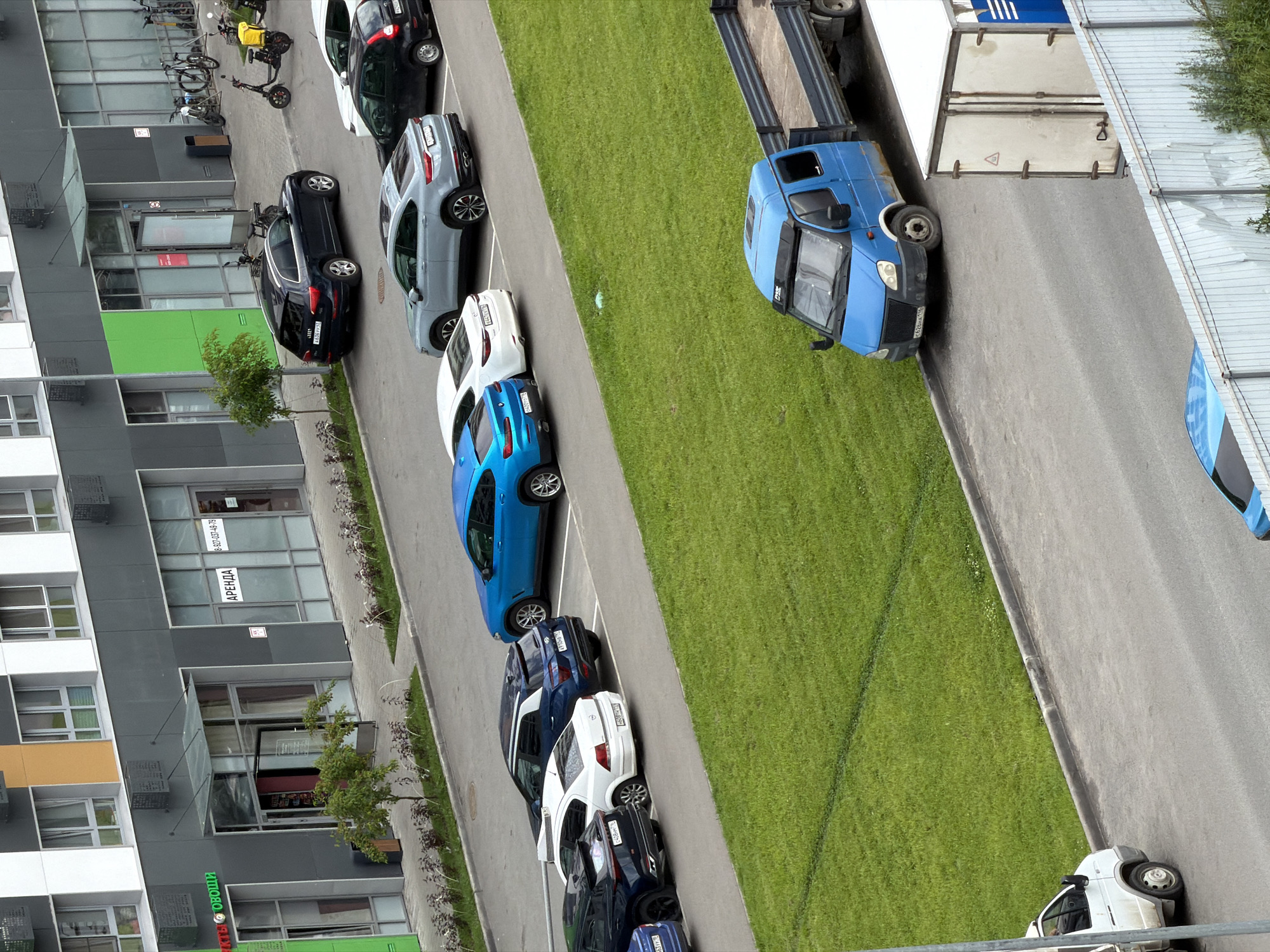 The image depicts an outdoor scene of an urban environment, likely a residential or commercial area, with a focus on parked vehicles and buildings. The foreground shows a gray asphalt road with a white van parked on the left and a blue flatbed truck parked in the middle. Beyond the road, there is a strip of green grass and then a row of parked cars, including a blue BMW, a white sedan, and several other dark-colored vehicles. In the background, a modern building complex is visible, featuring a facade with gray and white elements, large glass windows, and green accents. Various businesses or offices appear to be located on the ground floor, indicated by signage like "АРЕНДА" (rent) and a phone number. Bicycles and electric scooters are also parked near the building entrance, suggesting pedestrian or micro-mobility activity. The overall impression is one of a well-maintained urban setting with functional infrastructure for transportation and commerce.