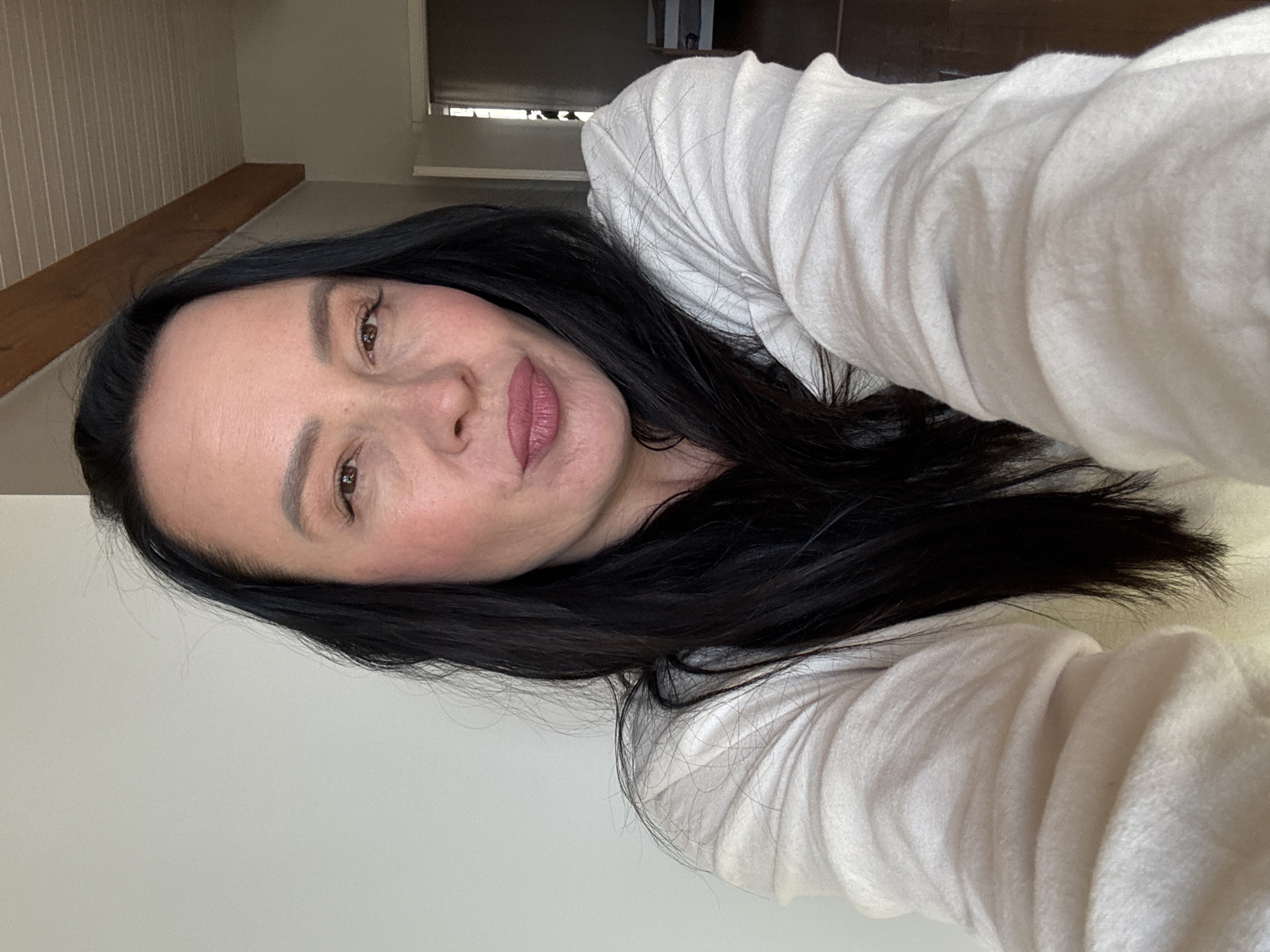 A selfie photograph shows a middle-aged woman with fair skin, dark hair, and lipstick. She is wearing a light-colored, long-sleeved garment. The background includes a wall, wooden beam, and a window, suggesting an interior space. The location, Chaikovsky, Russia, is not discernible from the image itself.