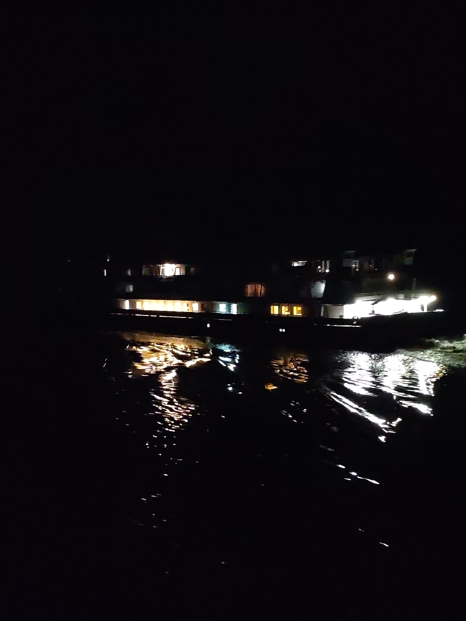 At night in Глебово, Russia, a multi-story passenger vessel is on the water. Lights illuminate the boat's exterior and reflect on the water's surface. The surrounding area is dark.
