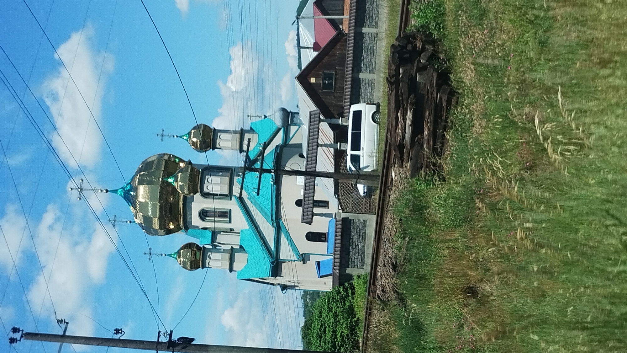 This media file depicts a picturesque scene in Teresva, Ukraine, featuring an Eastern Orthodox church with striking golden domes and a vibrant turquoise roof. The church is situated next to a railway track, with piles of wood and lush green grass flanking the tracks, suggesting a rural or semi-rural setting. A white van is parked near the church, and the sky above is a brilliant blue with scattered white clouds, indicating pleasant weather, likely during the daytime. Overhead power lines and a utility pole are visible in the foreground, adding to the sense of a lived-in landscape. The overall impression is one of tranquility and beauty, with the sunlight reflecting off the church's golden domes. There is no visible text in the image.