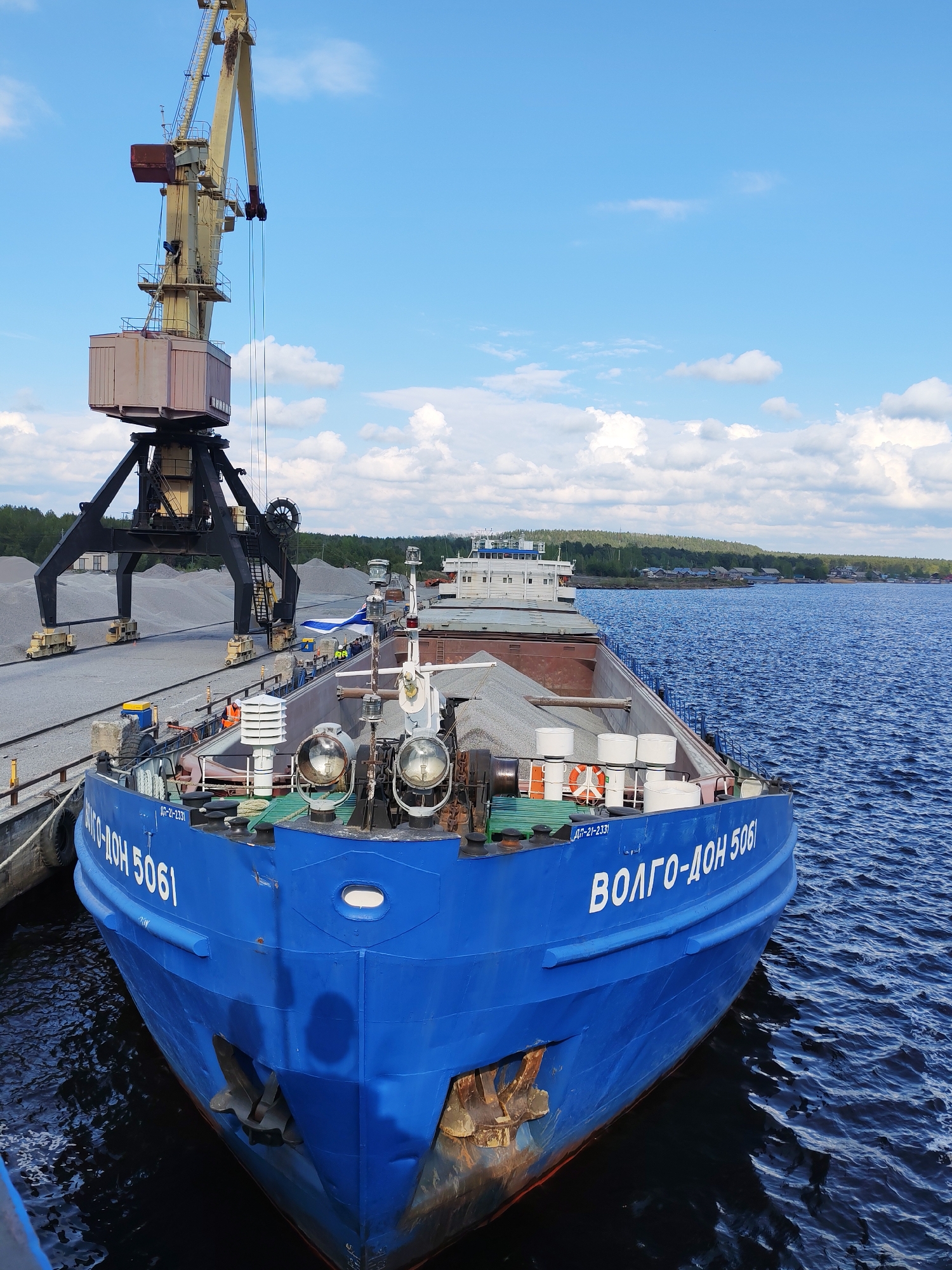 The image shows a large blue river barge named "VOLGO-DON 5061" docked at a port in Shal'skii, Russia. A yellow crane with a gray control cabin is positioned to the left of the barge, near piles of gray gravel. The barge is loaded with gravel. Life preservers are visible along the edges of the boat. The water is a dark blue with gentle ripples. A treeline and village buildings are visible across the water. The sky is blue with scattered white clouds. Several people are working on or near the barge, wearing safety vests.